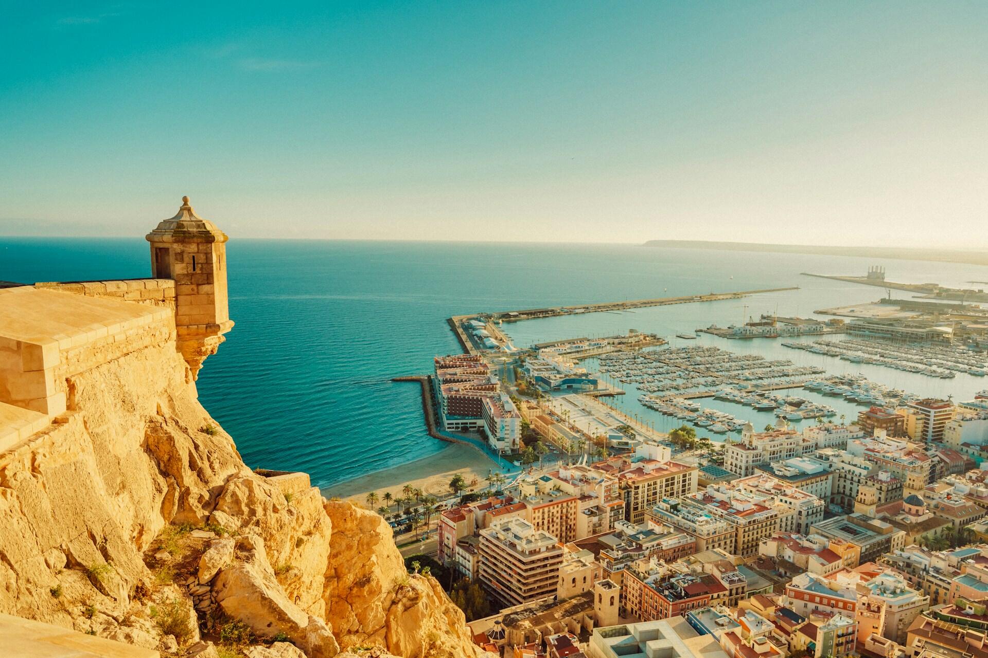 Aerial view of a coastal town with colorful buildings, a marina filled with boats, and a cliffside castle tower overlooking the sea.