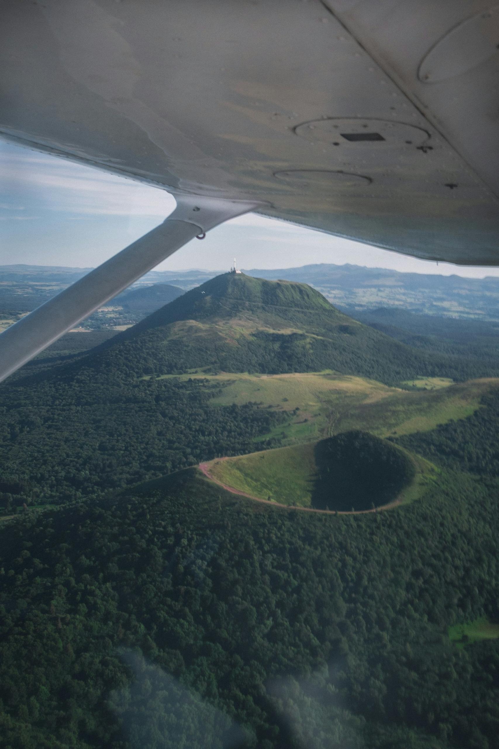 An airplane flies over a volcanic crater covered in greenery during the day. 