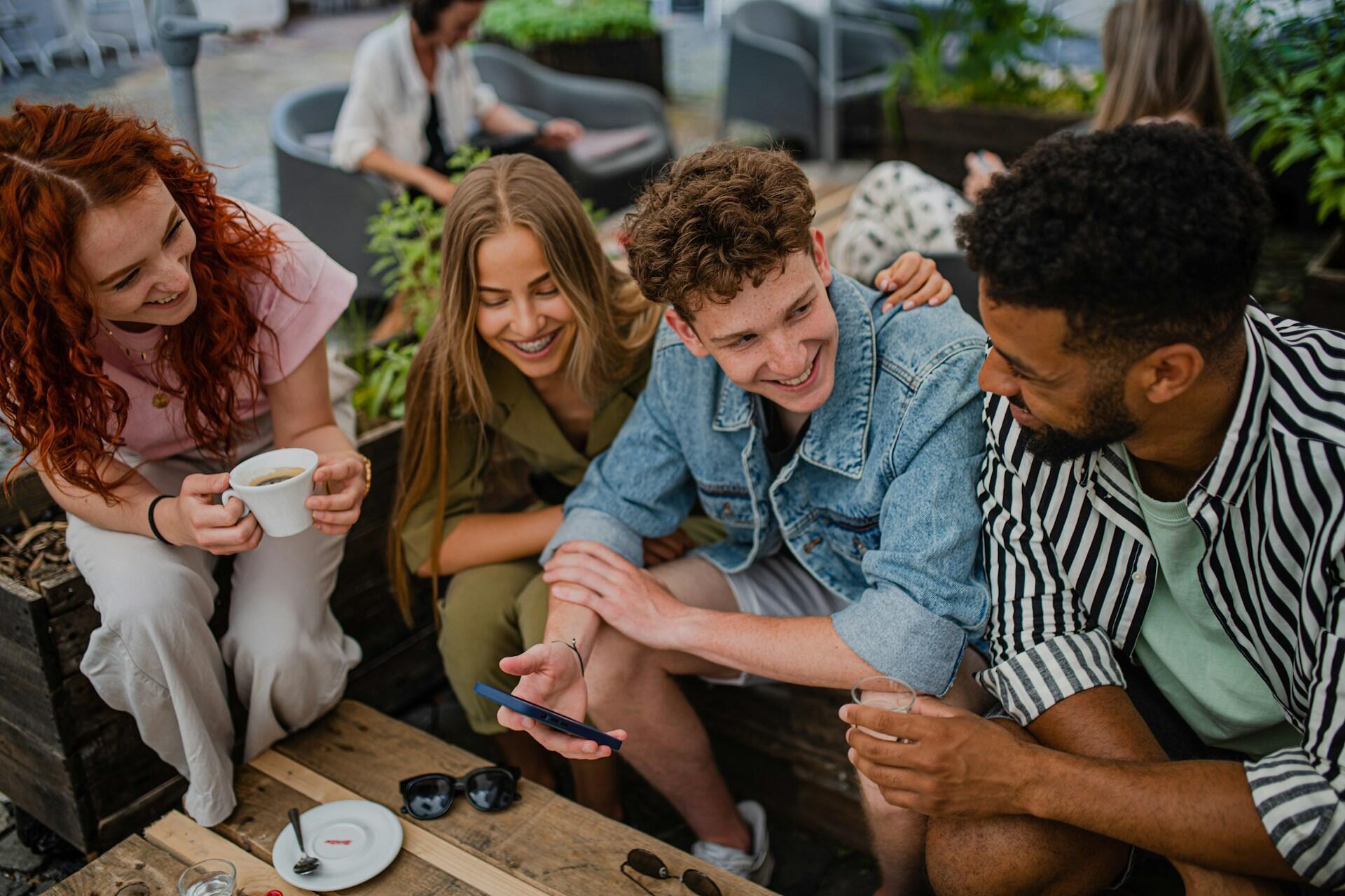 A group of young people sitting outside around a wooden table, laughing and drinking coffees.