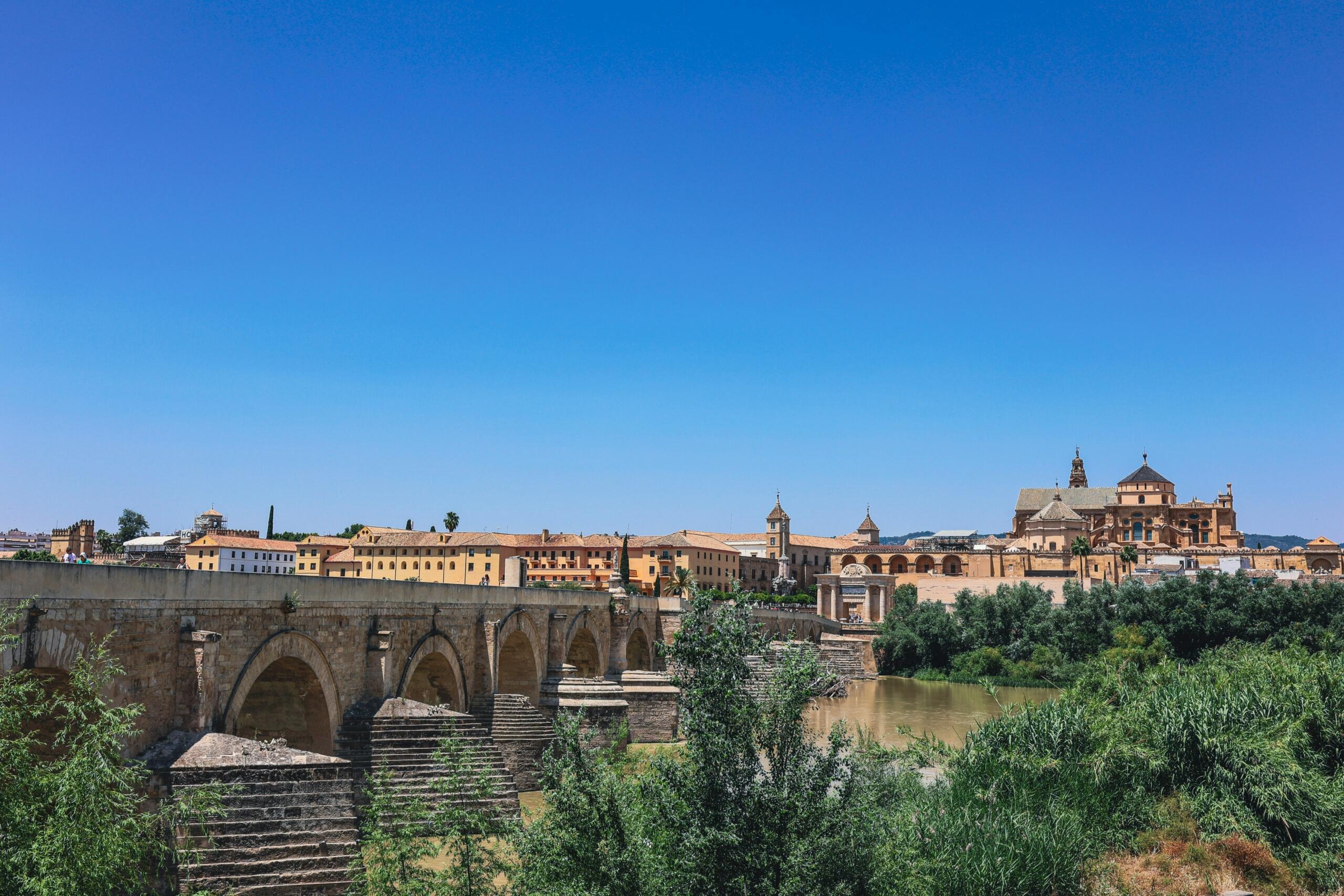 A scenic view of a historic stone bridge over a river, with colorful buildings and greenery under a clear blue sky.