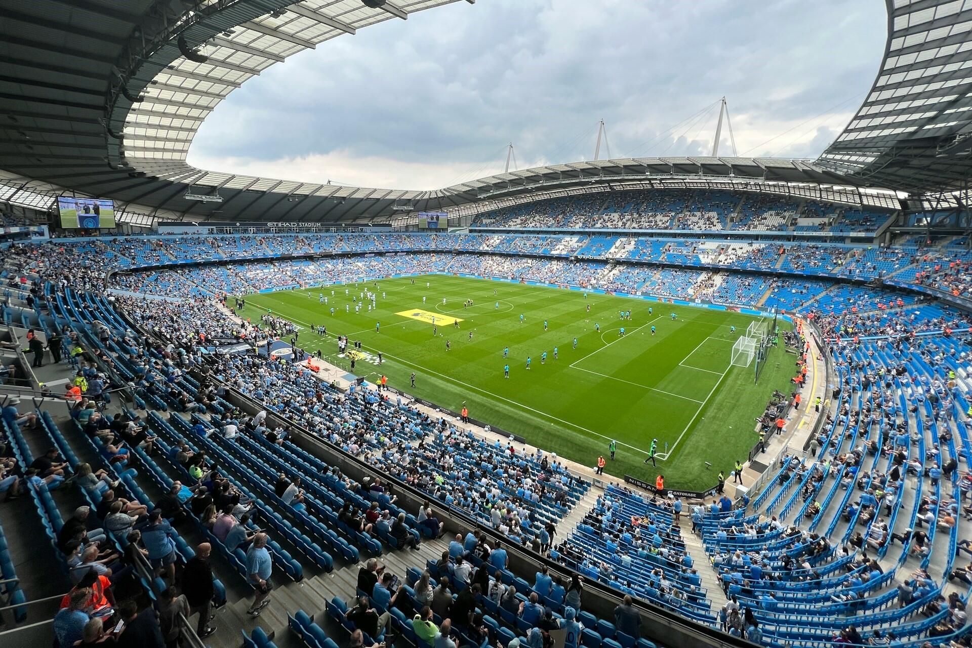 Wide view inside Etihad Stadium with blue seats, a football pitch and spectators in the stands