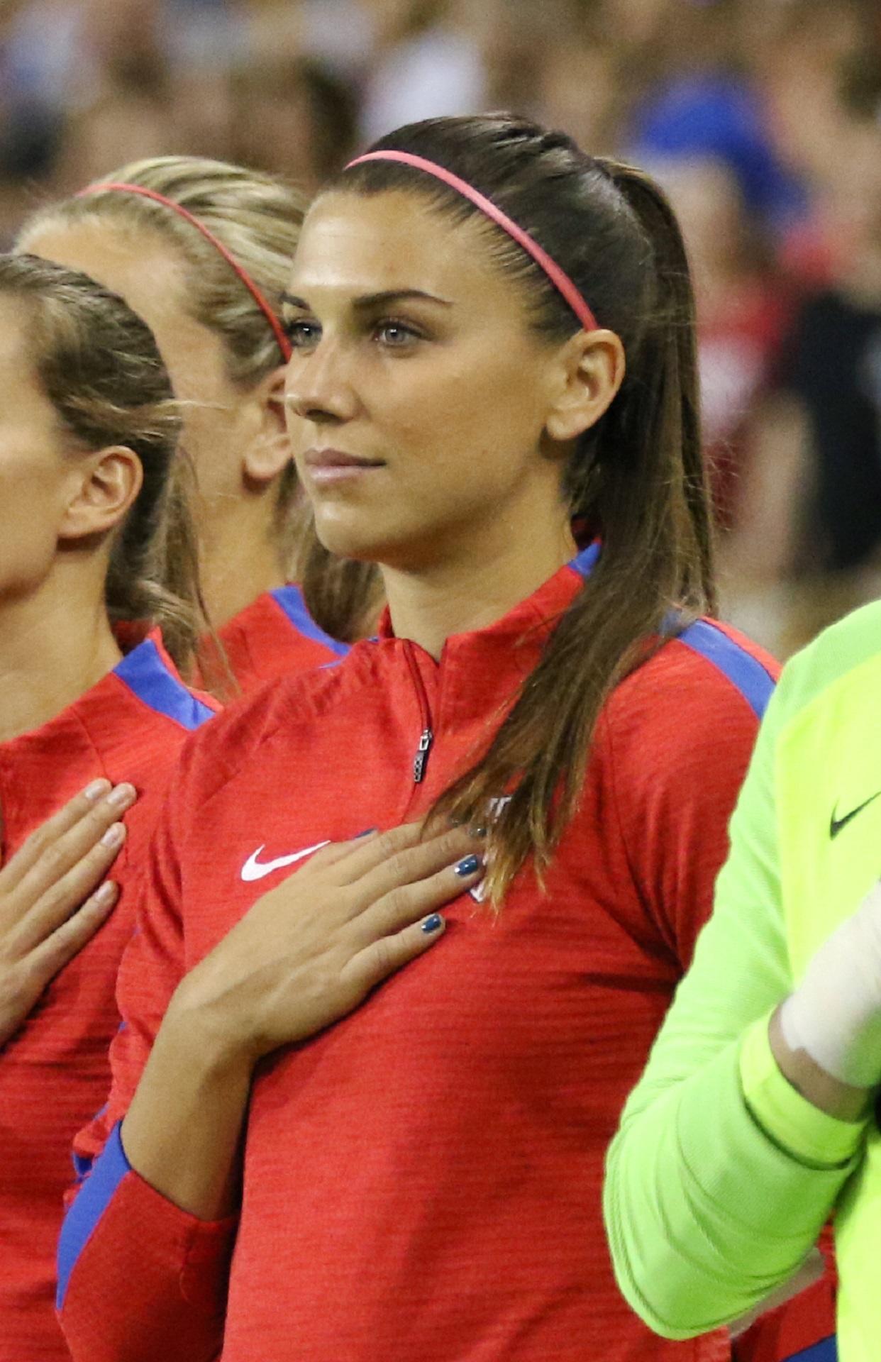 A person in a red top with their hair pulled back and hand over heart.