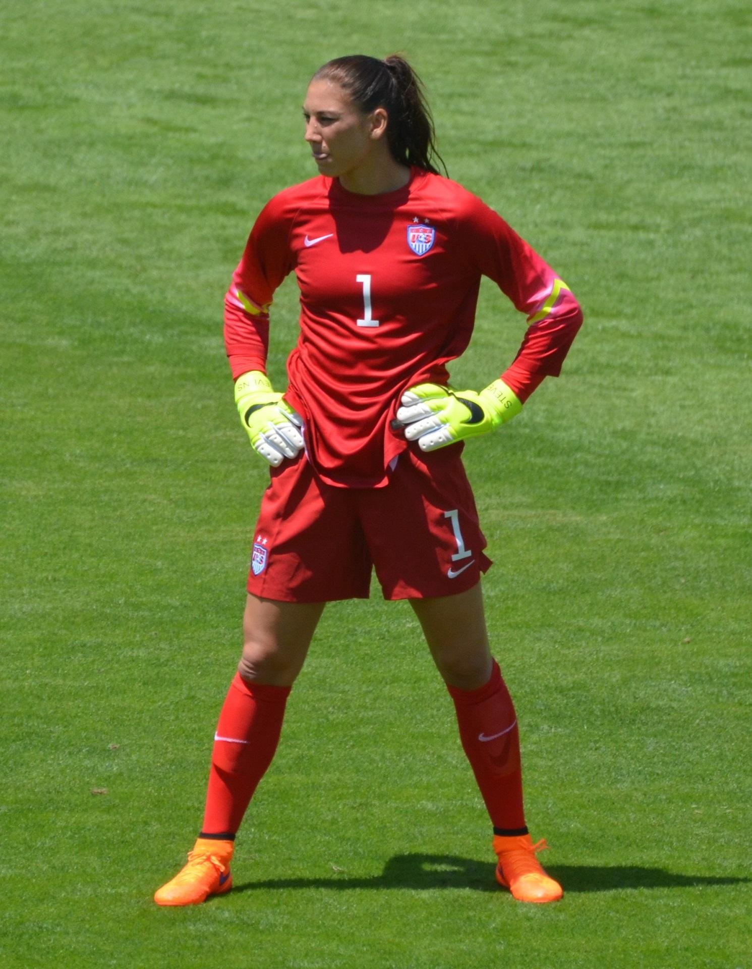 A person in a red football uniform stands on the pitch with arms akimbo.
