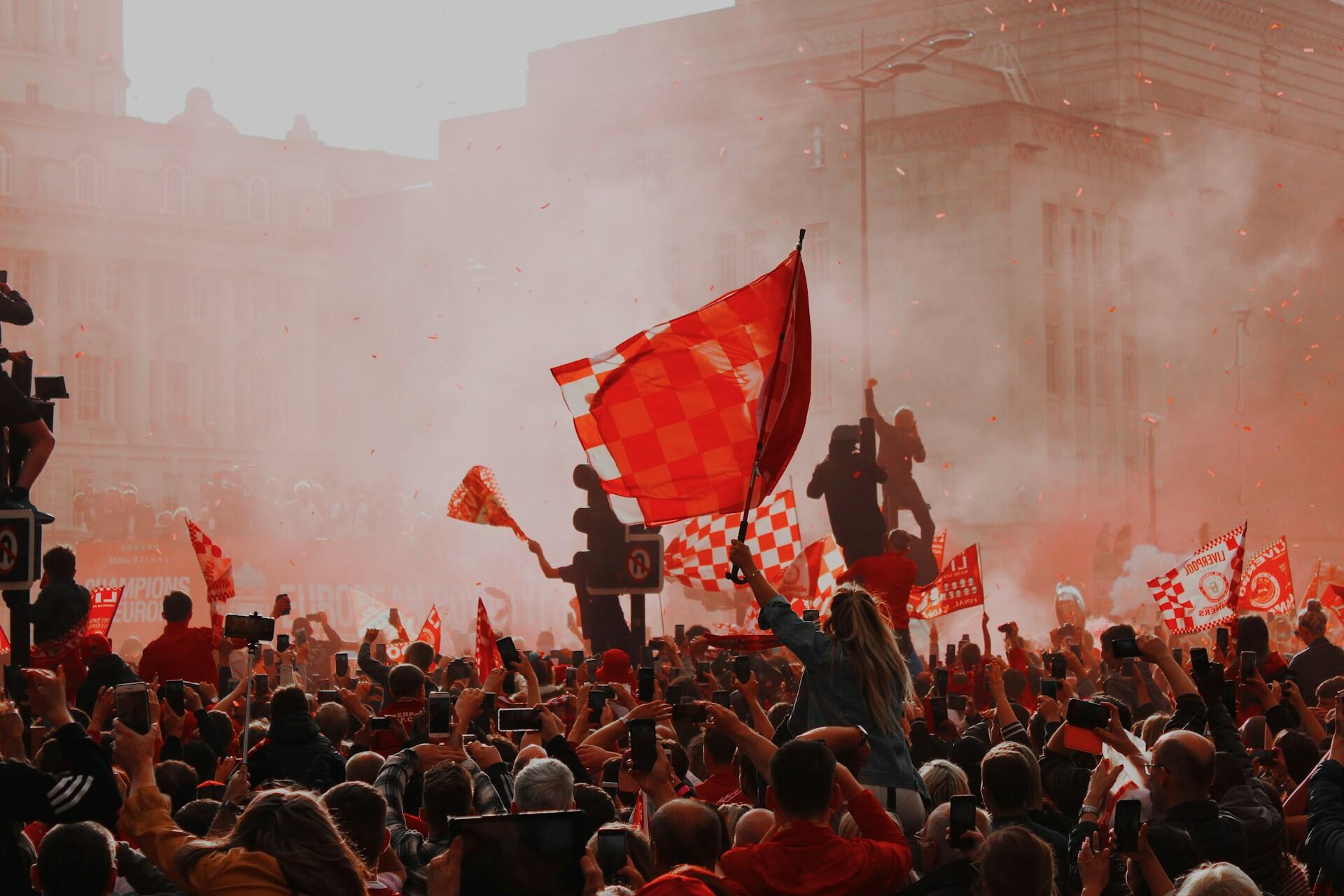 Crowd of supporters holding red flags and phones during a celebration in a city street filled with smoke