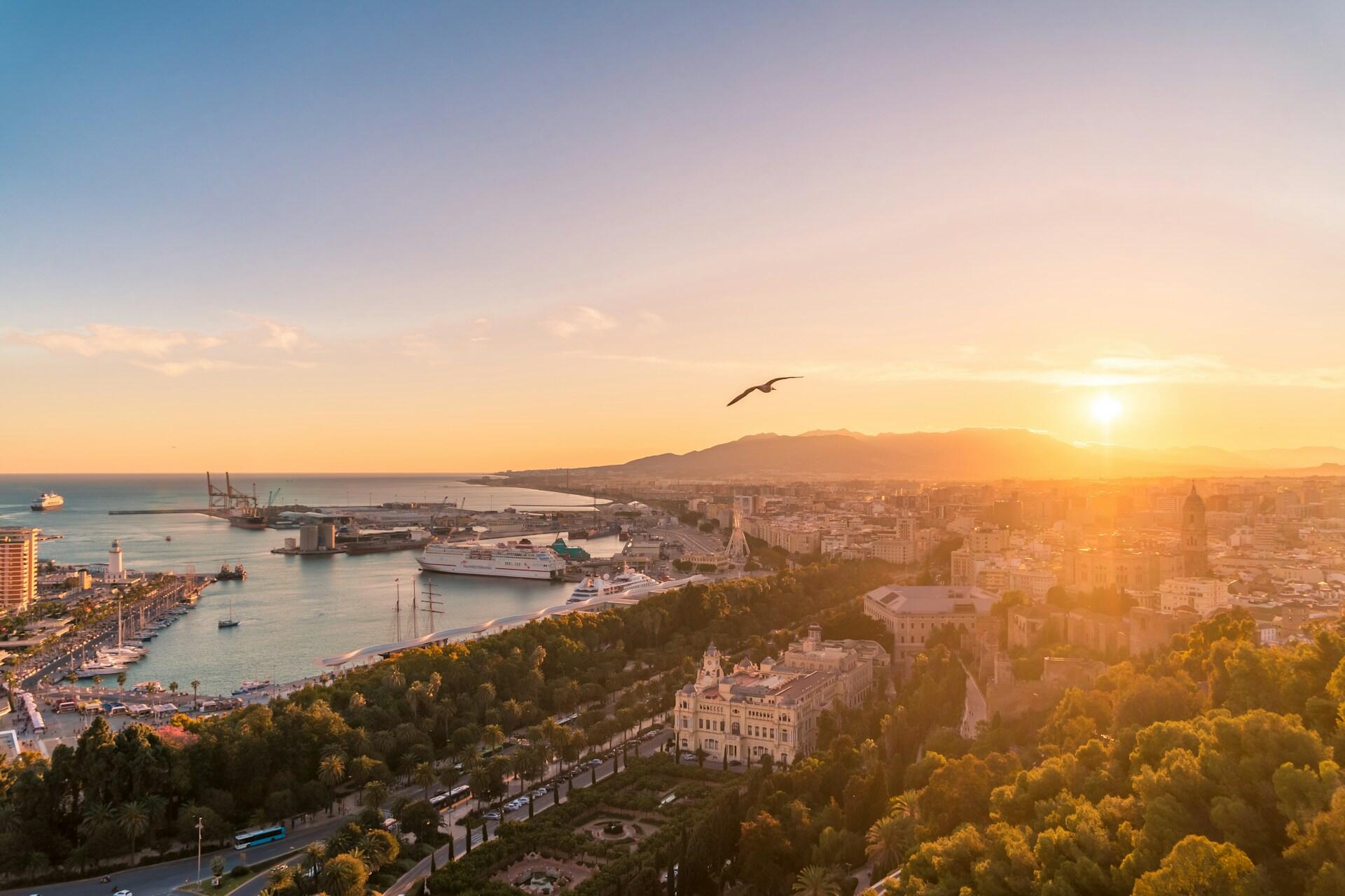A stunning sunset over a coastal city, featuring a vibrant harbor with ships, palm trees, and mountains in the background.