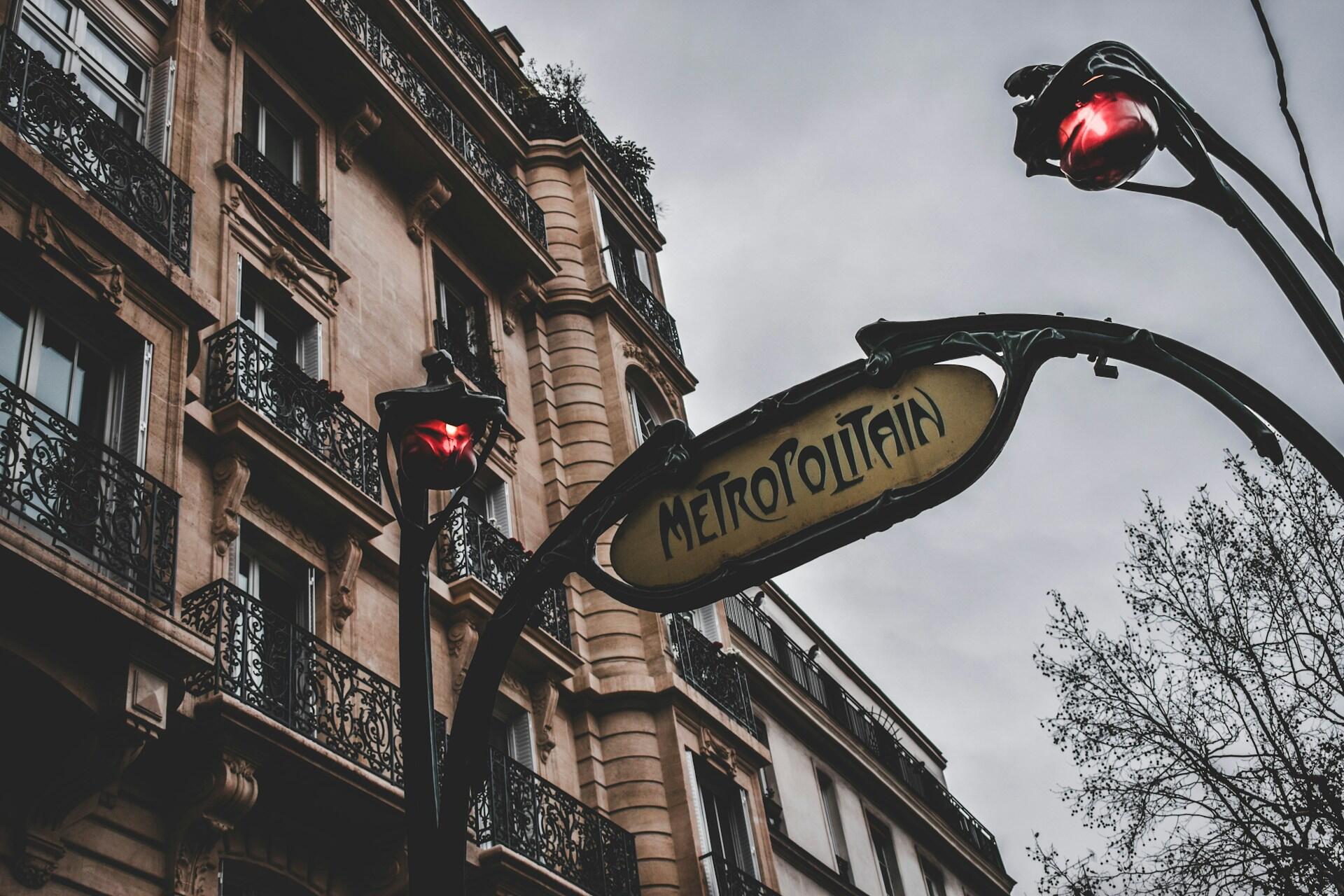 A metro sign mounted on iron poles between two lights in front of a stone building.