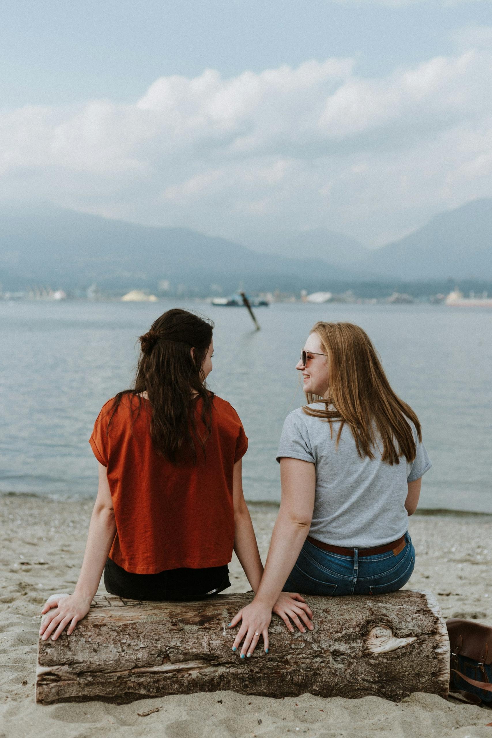 Two people sit on a log at the beach on a cloudy day.