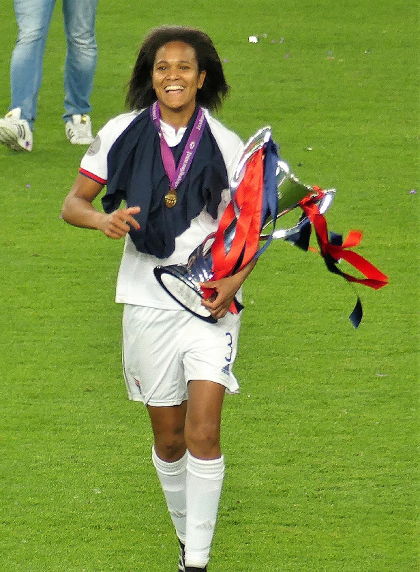 A woman wearing a white sports uniform carries a silver trophy.