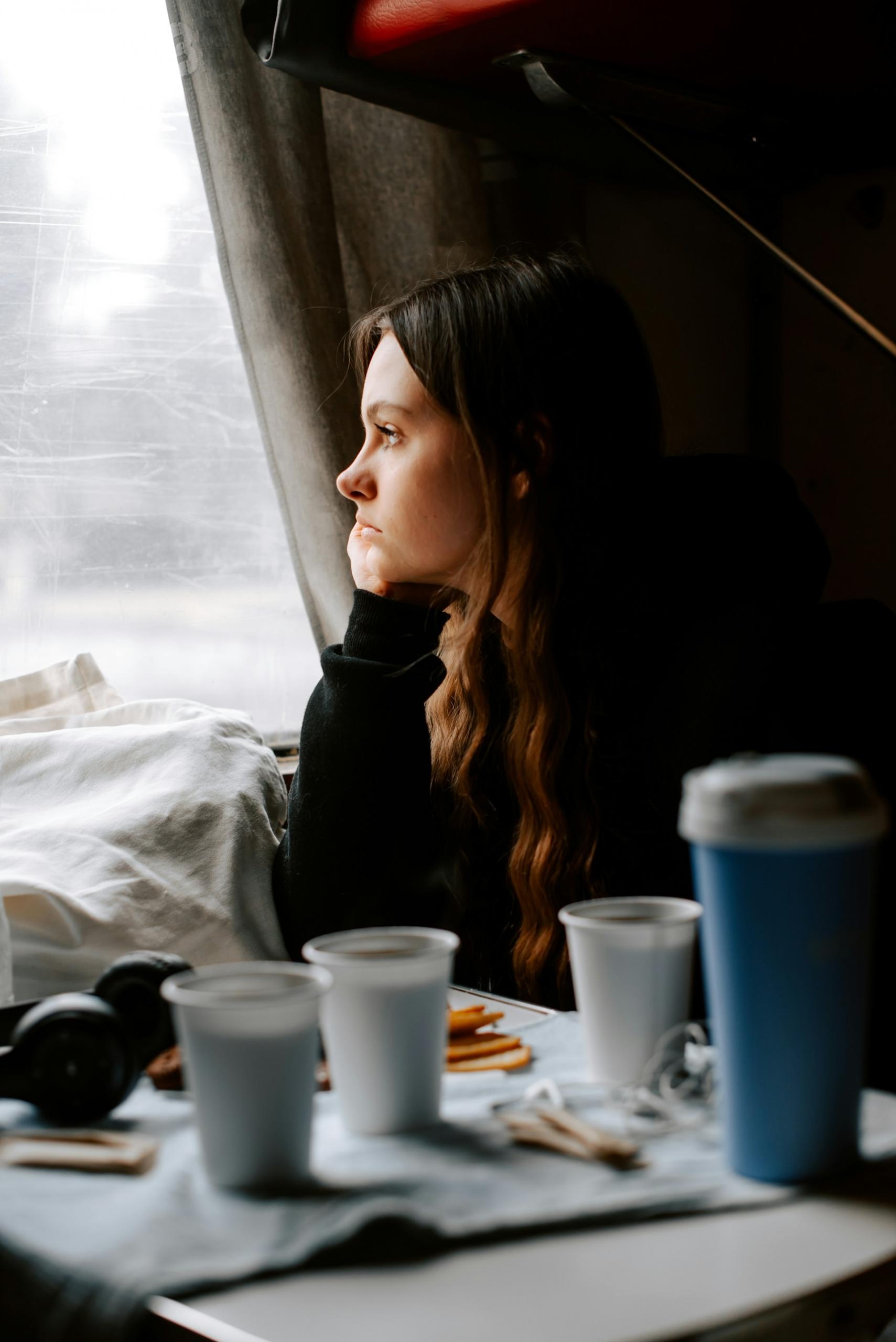 A woman looks out the window while sitting at a table with drinking cups on it. 