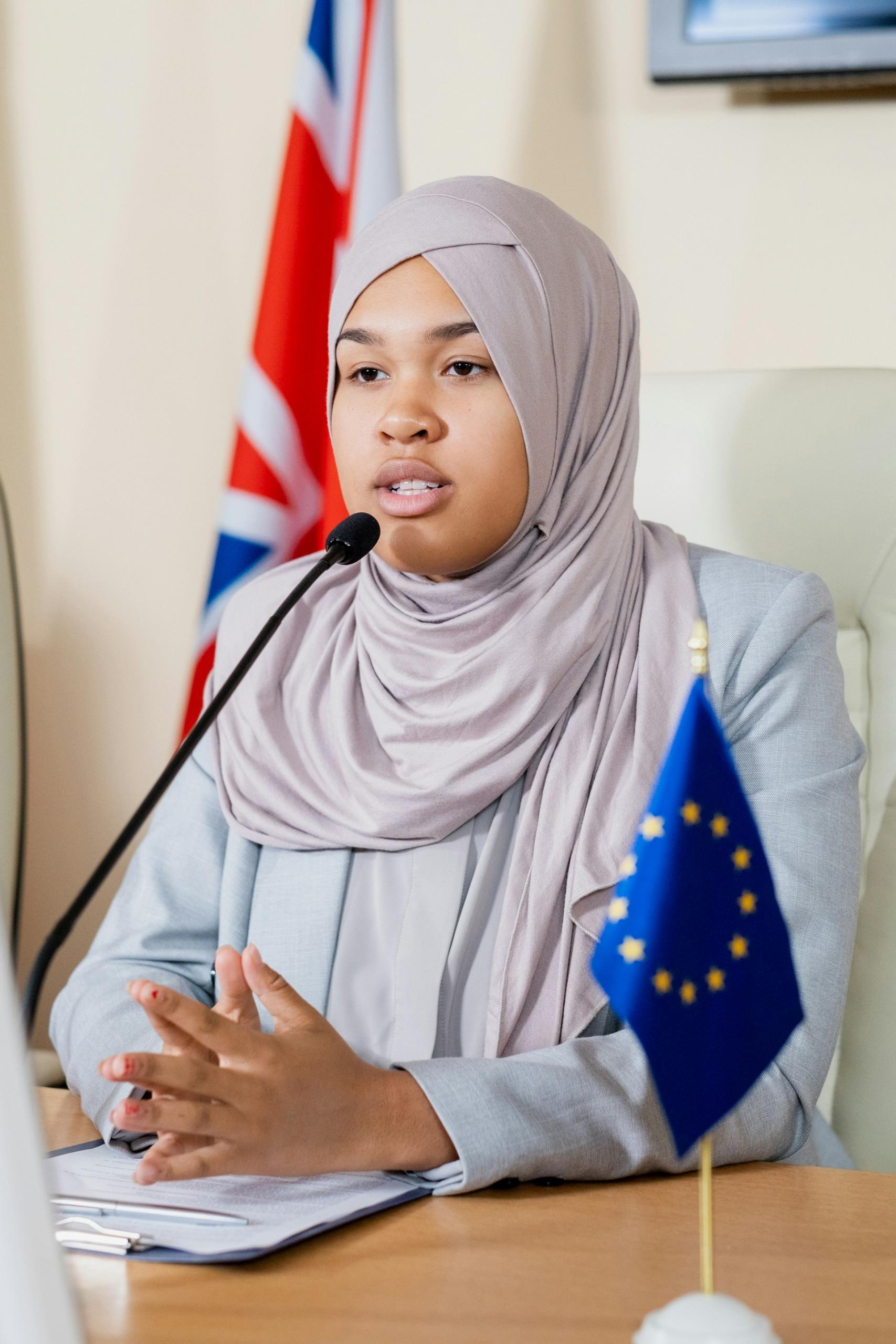 A woman wearing a hijab sits at a table with a small European Union flag on it, and a large UK flag in the background. 