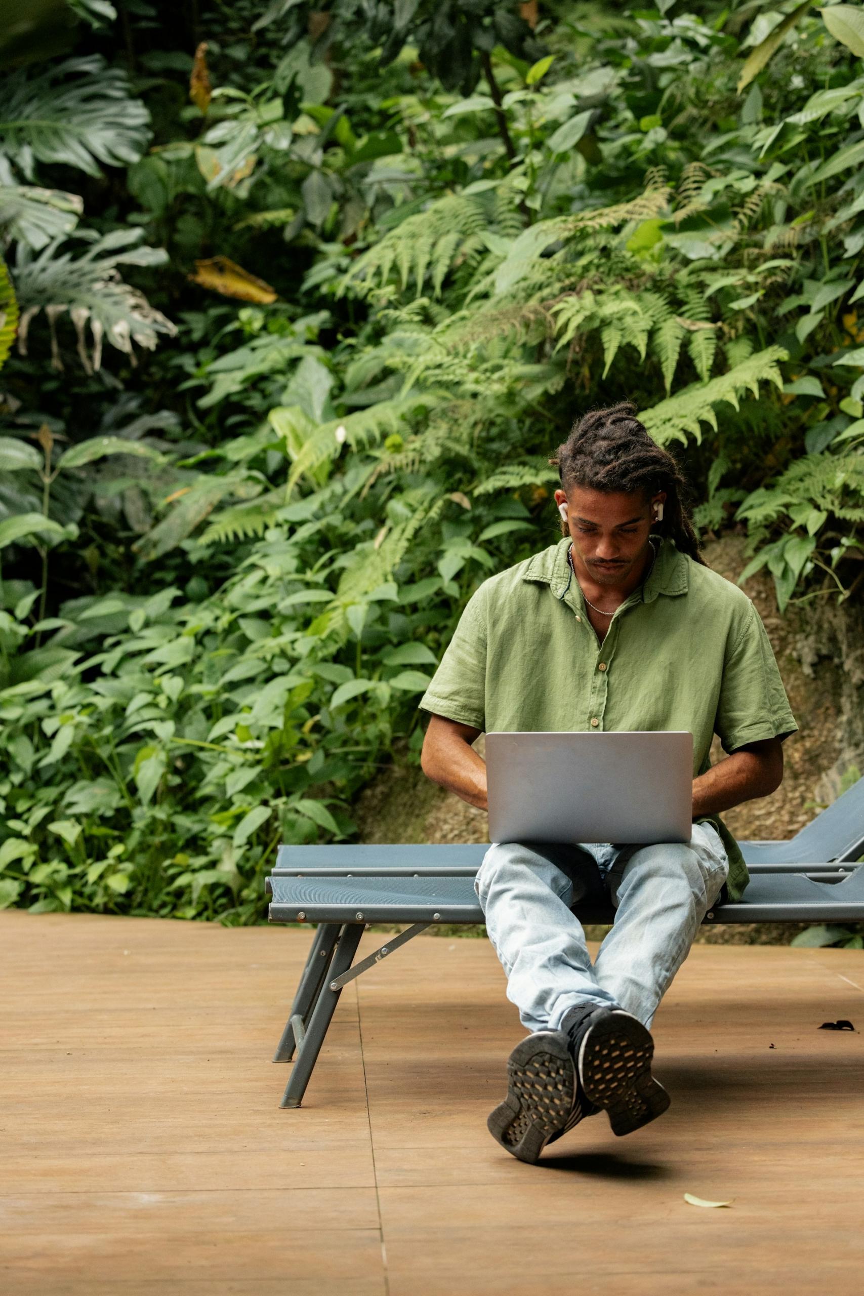 A man sitting on a bench outside working on a computer.