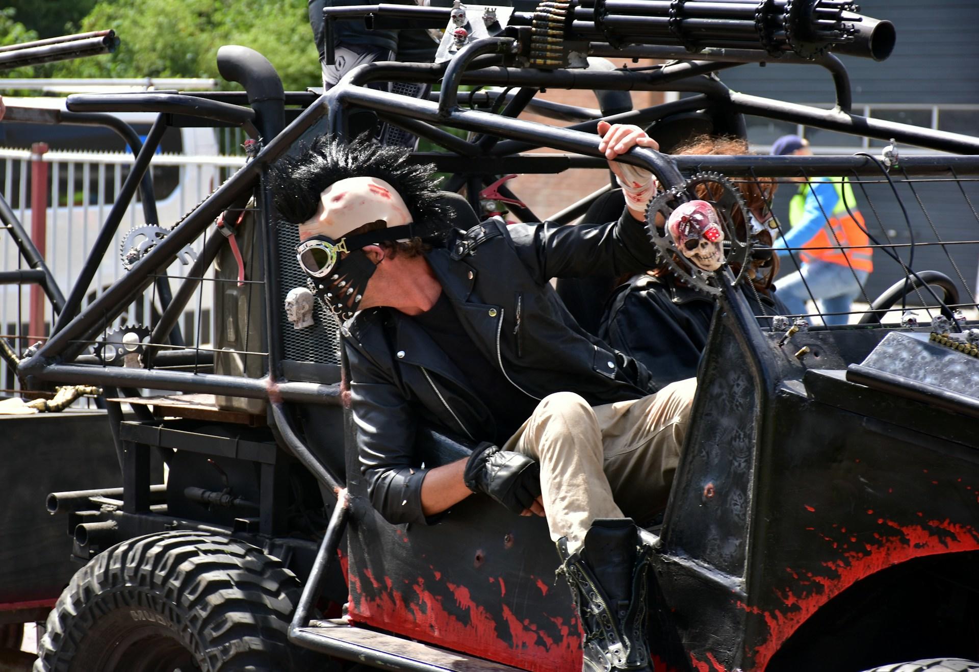 A man in costume, wearing a leather face mask with goggles, leans out of a black and red dune buggy.