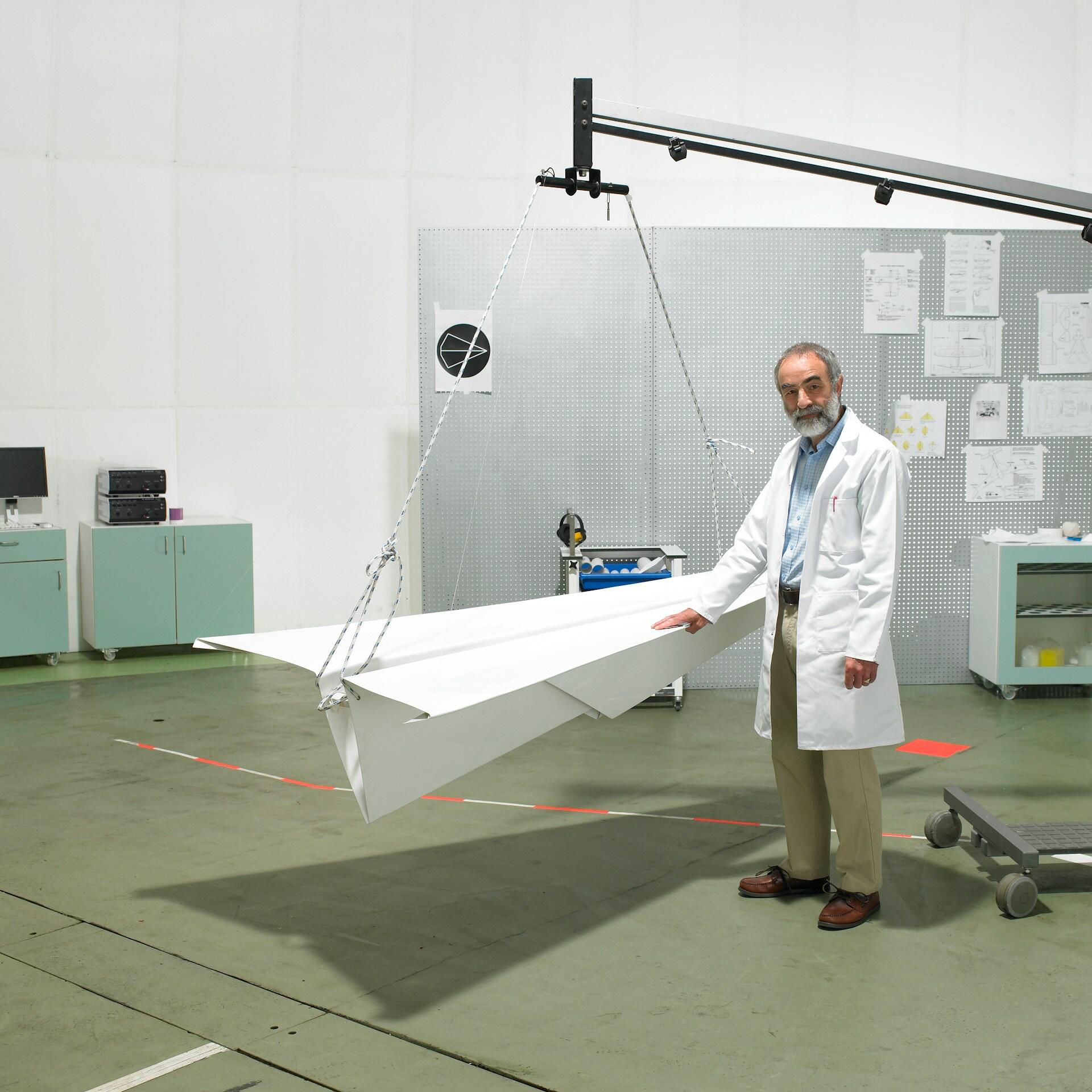 A man in a white labcoat stands next to a giant paper airplane suspended by a crane. 