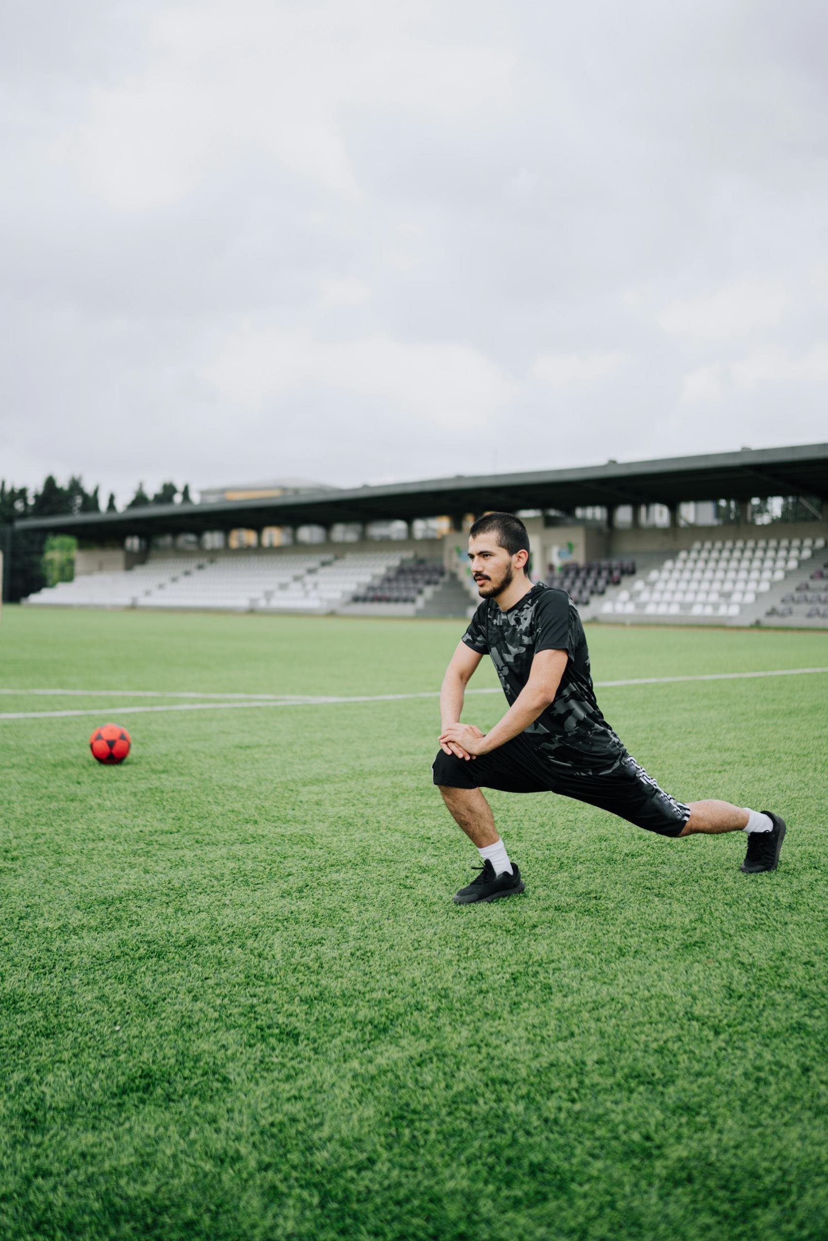 A man wearing sports clothing lunges on a soccer pitch on a cloudy day.
