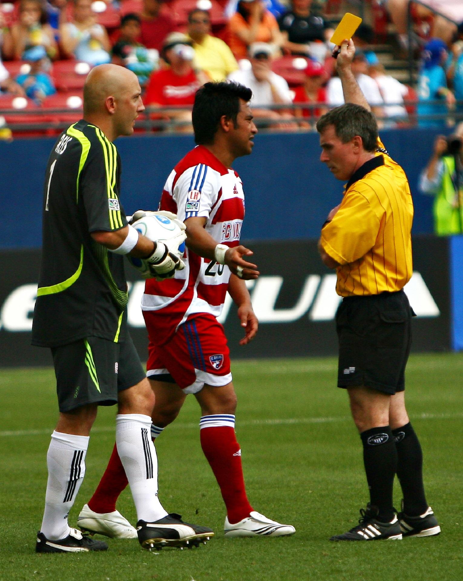A man in a yellow jersey confronts two players on a soccer pitch. 