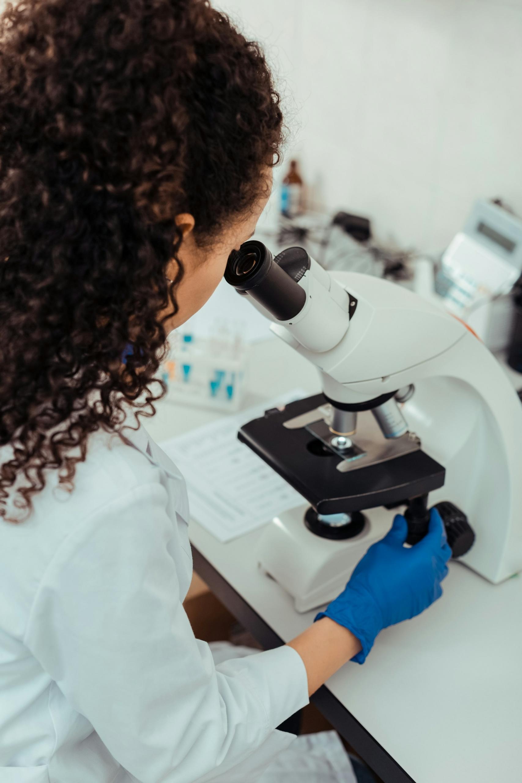 A woman wearing a labcoat and blue gloves looks into a microscope.