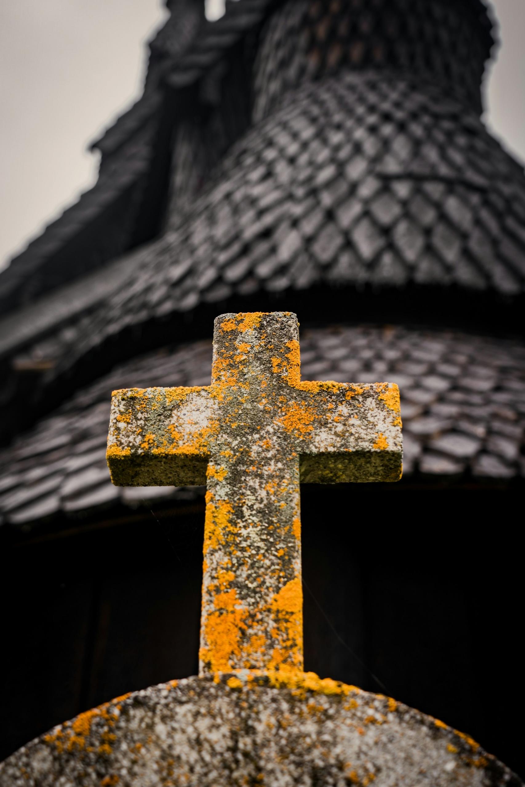 A stone cross covered with orange lichen, with a dark building behind it. 