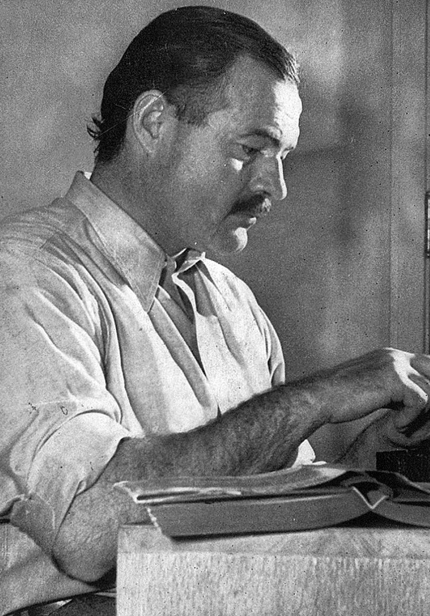 A black and white photo of Hemingway with a book open on the table.