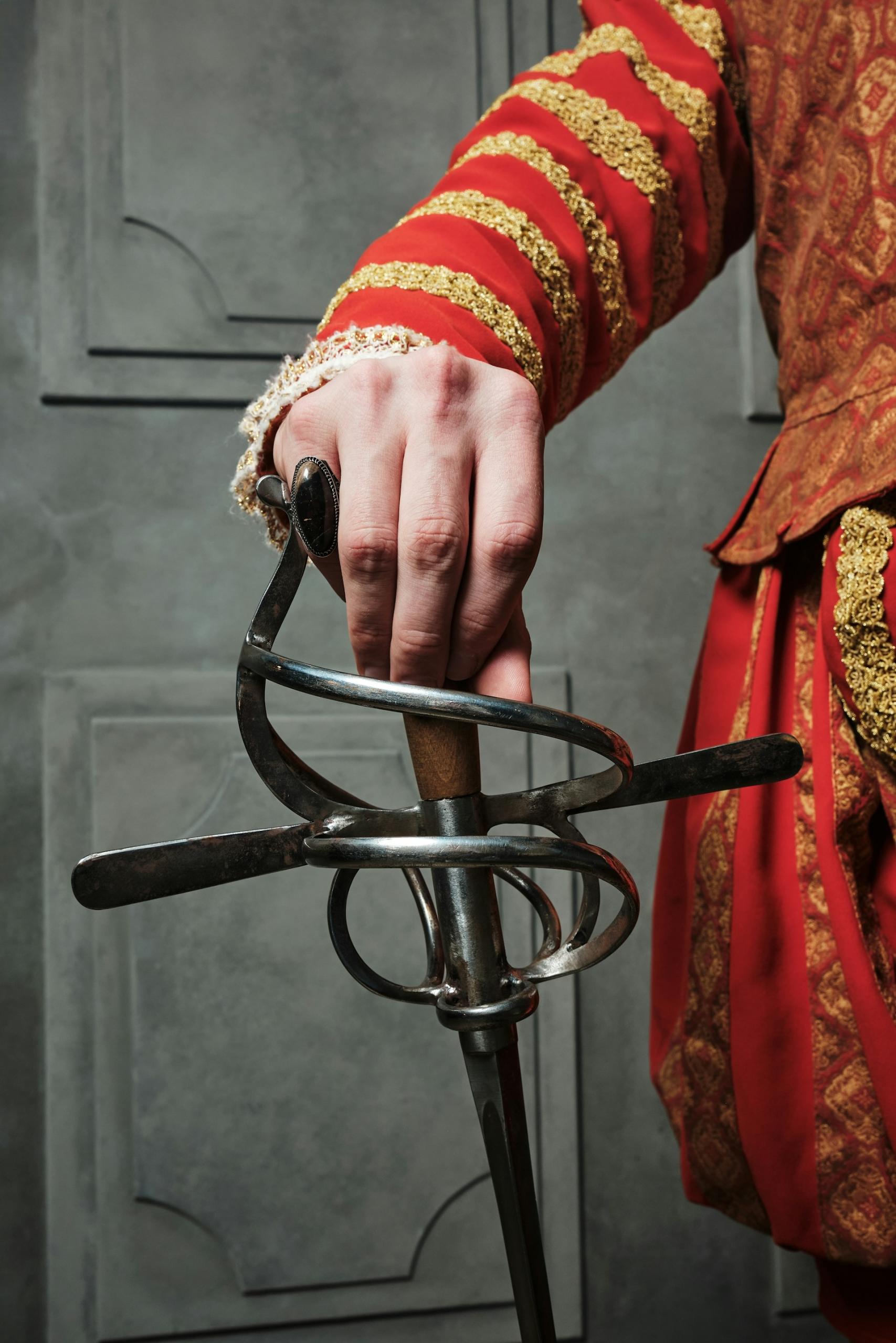 The arm and side of a man in red and gold clothing, with their hand on the butt of a sword. 