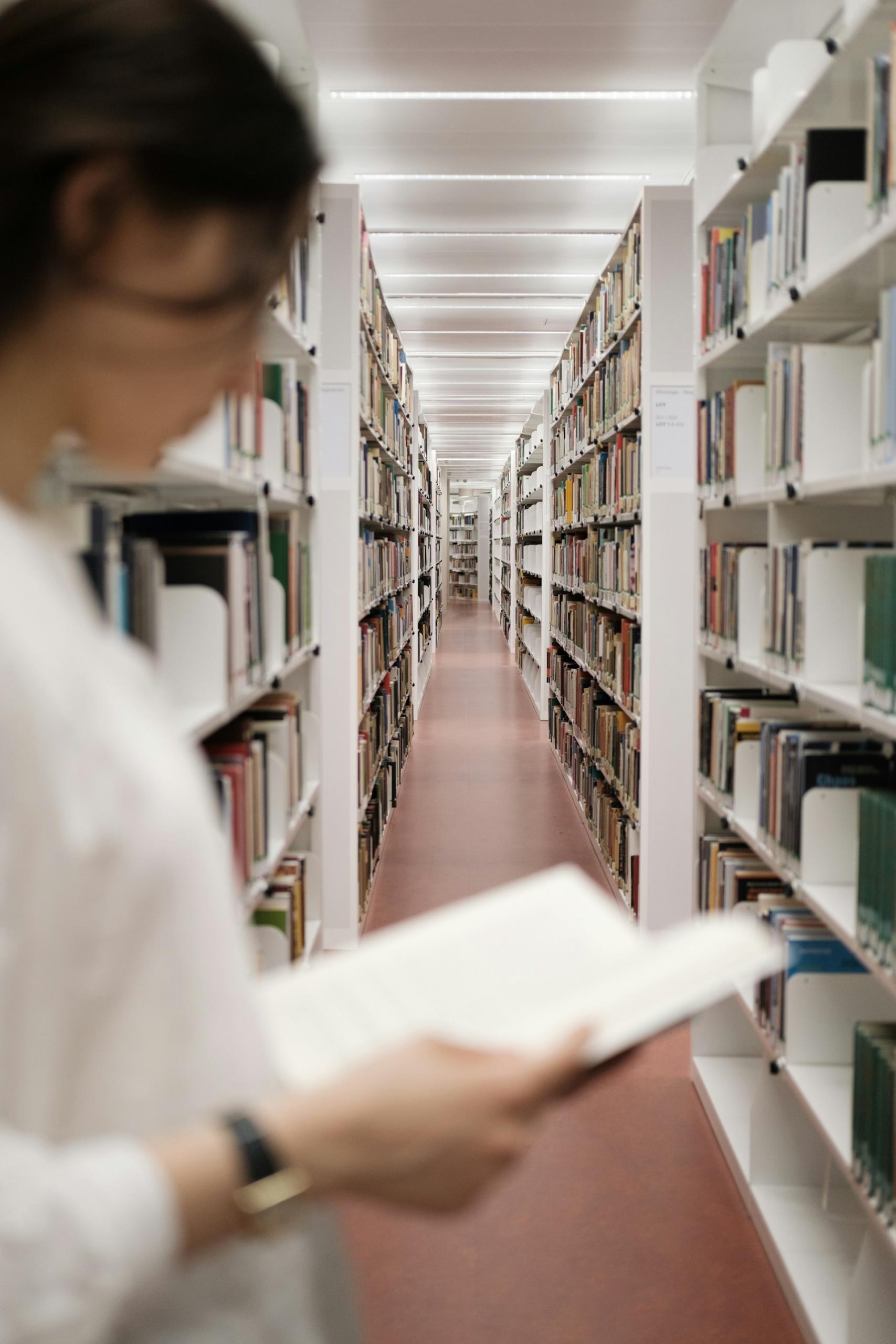 A person in a white shirt holds an open book in front of a library stack. 