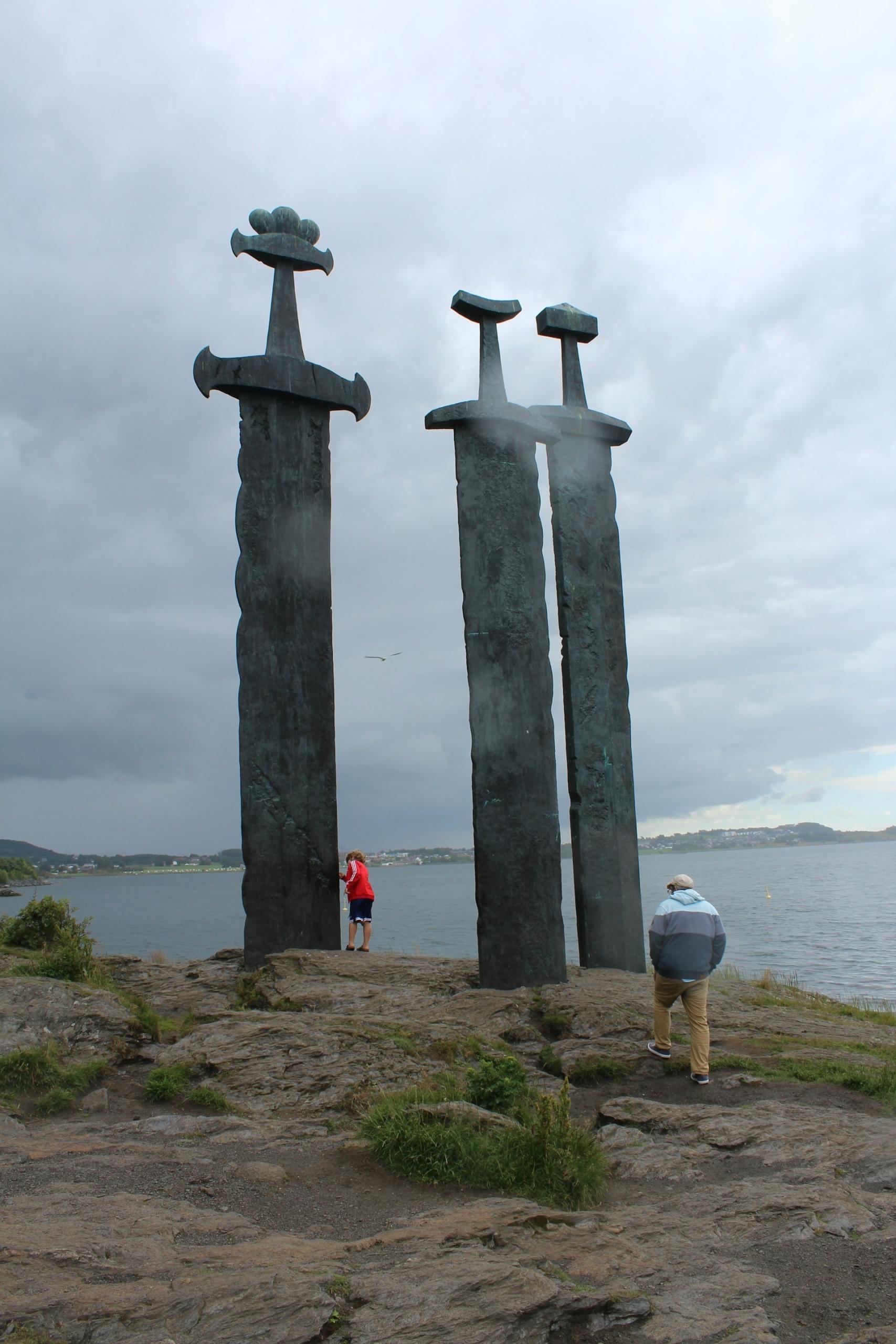 Three massive sword sculptures planted on a shore on a cloudy day.