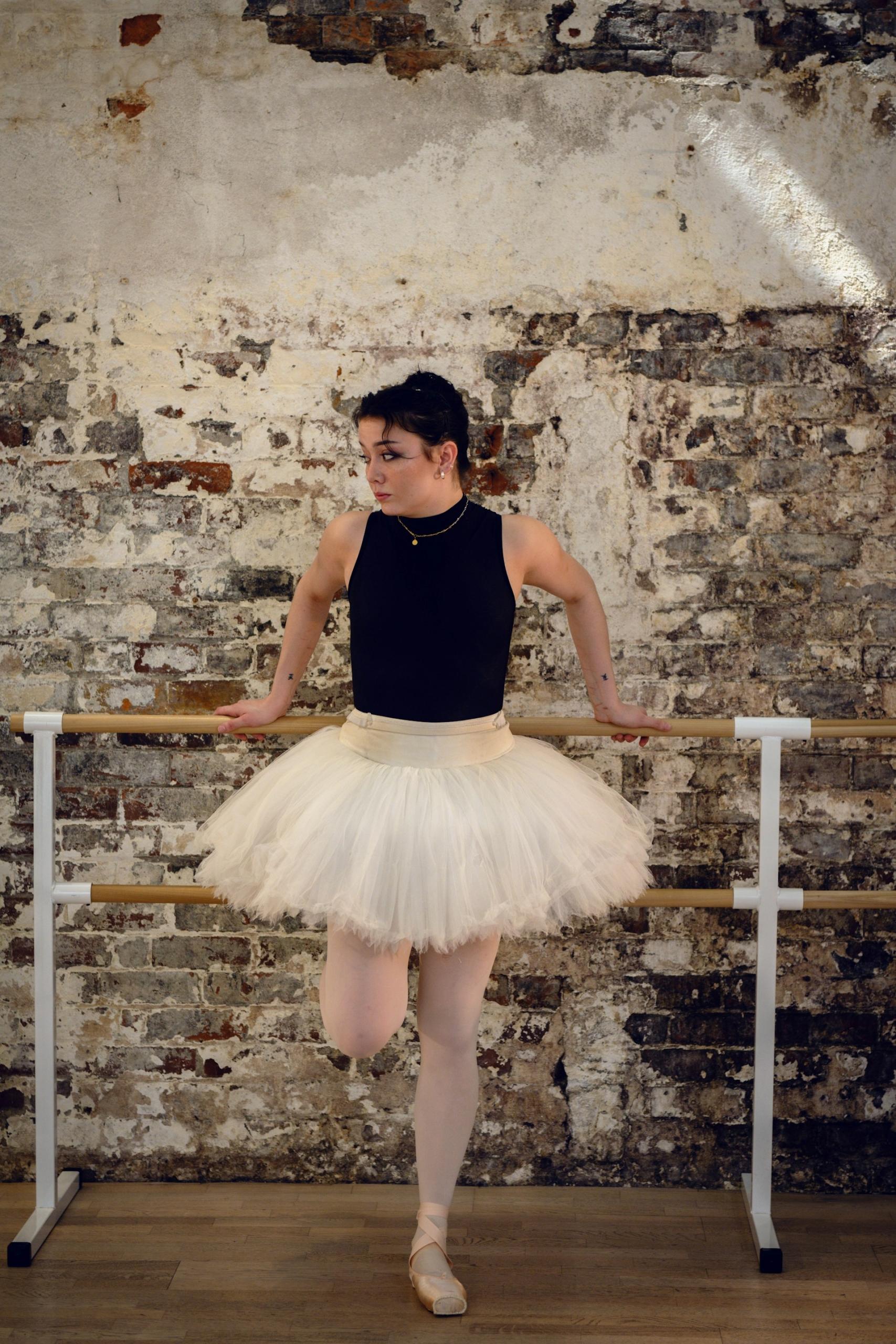 A woman in a pink tutu stands in front of a barre. 