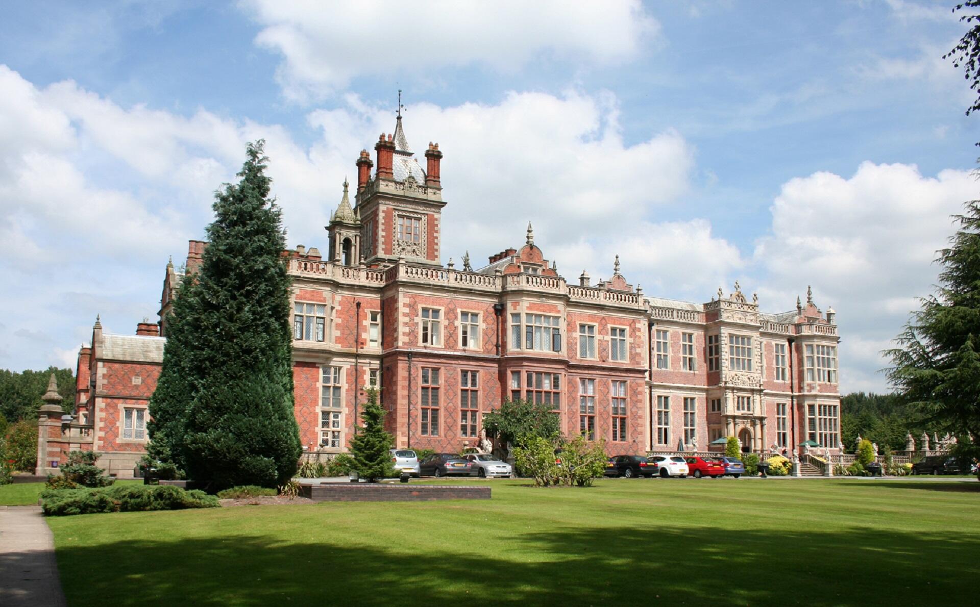 A large, ornate, red brick building behind an expansive green lawn on a cloudy day. 