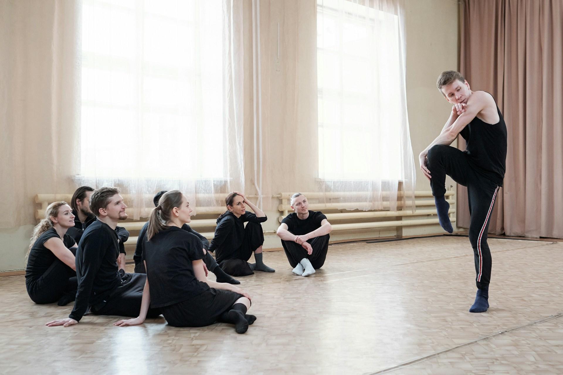 A man wearing black demonstrates a dance pose to black-clad students in a bright studio.