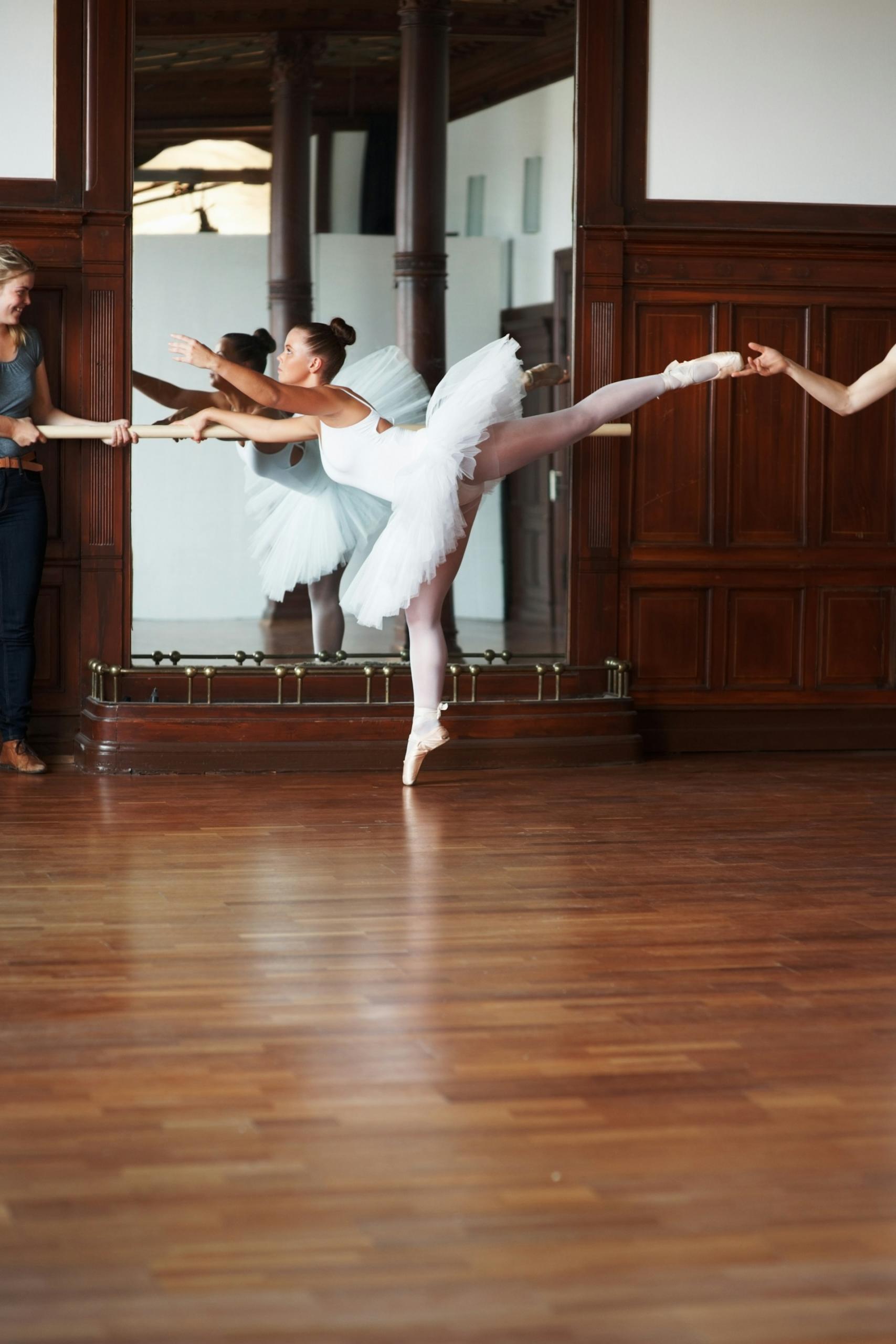 a woman in a tutu raises her leg while a person holds her foot.