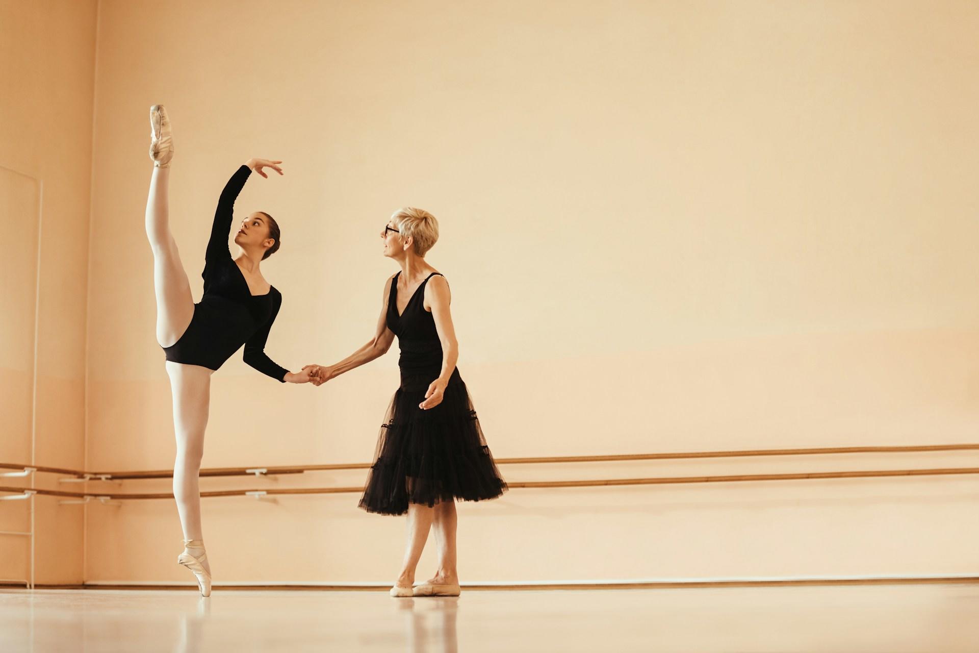 Two women wearing black dance in a studio with pale yellow walls.