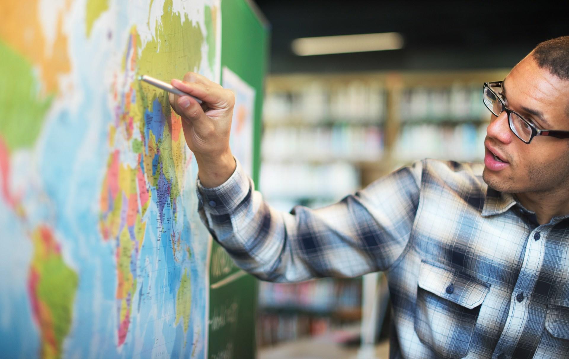 A man in a plaid shirt and glasses points to a map inside a library.