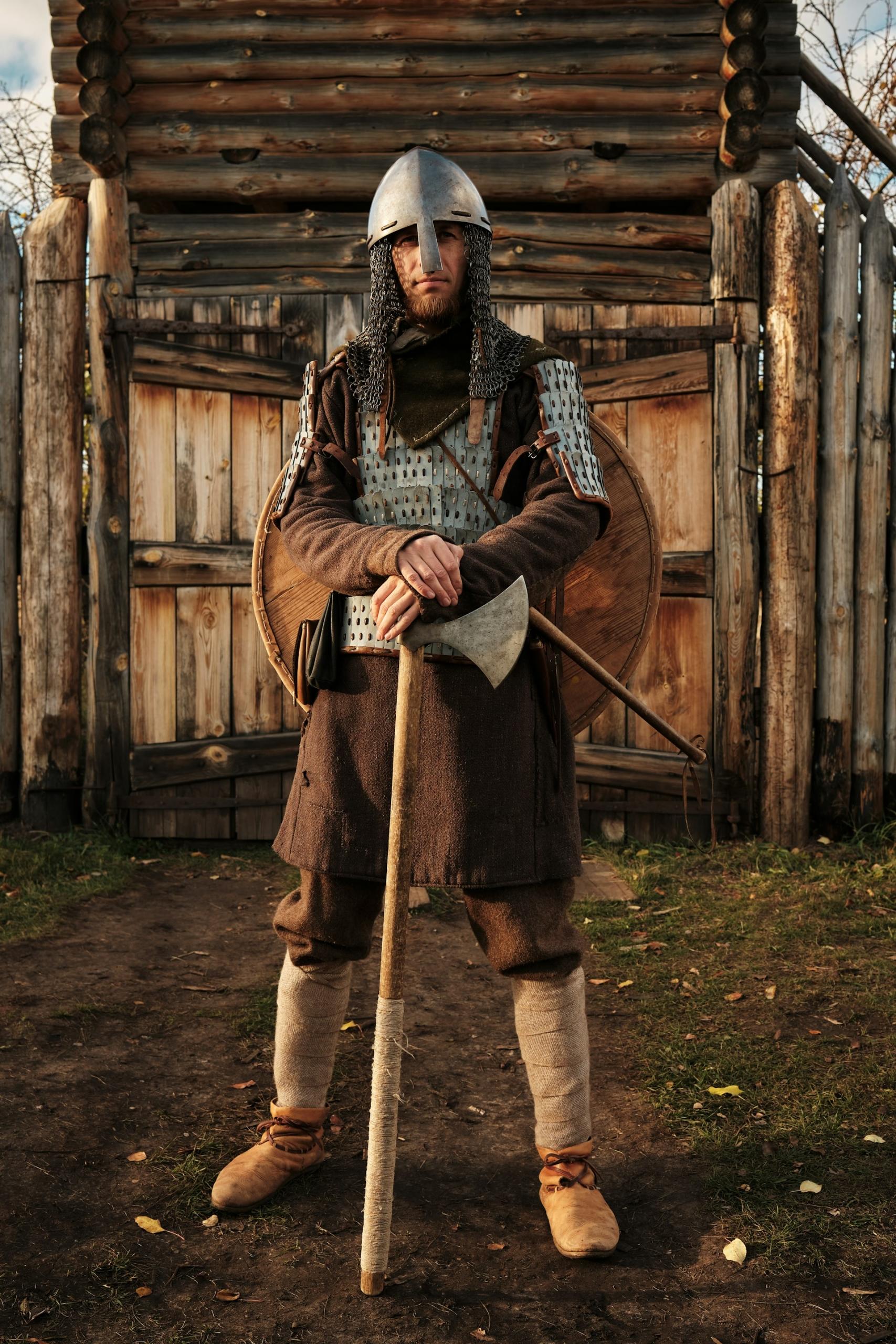 A man in Medieval costume stands in front of a wooden wall. 