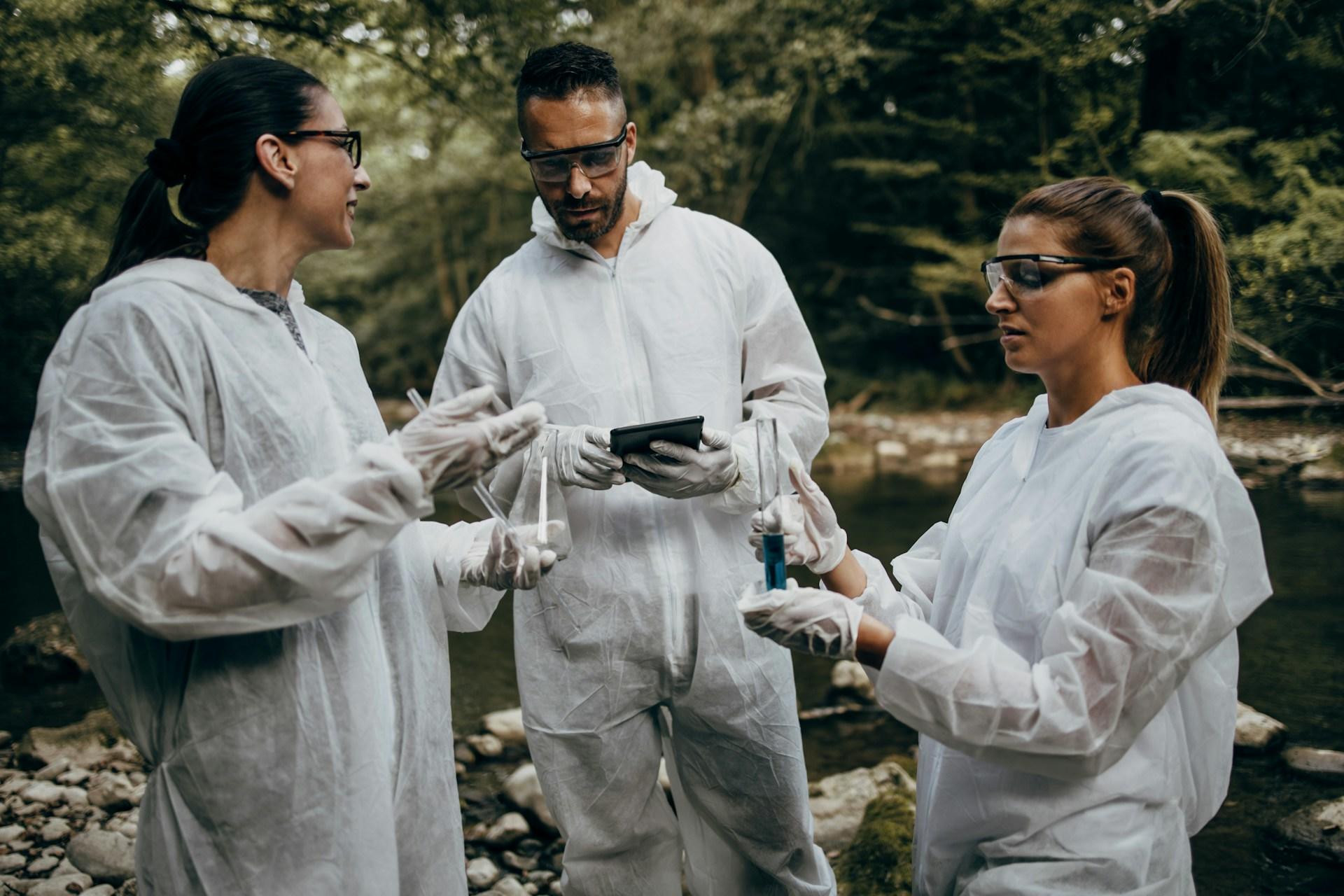 Three people in white safety suits stand together in a nature environment. 