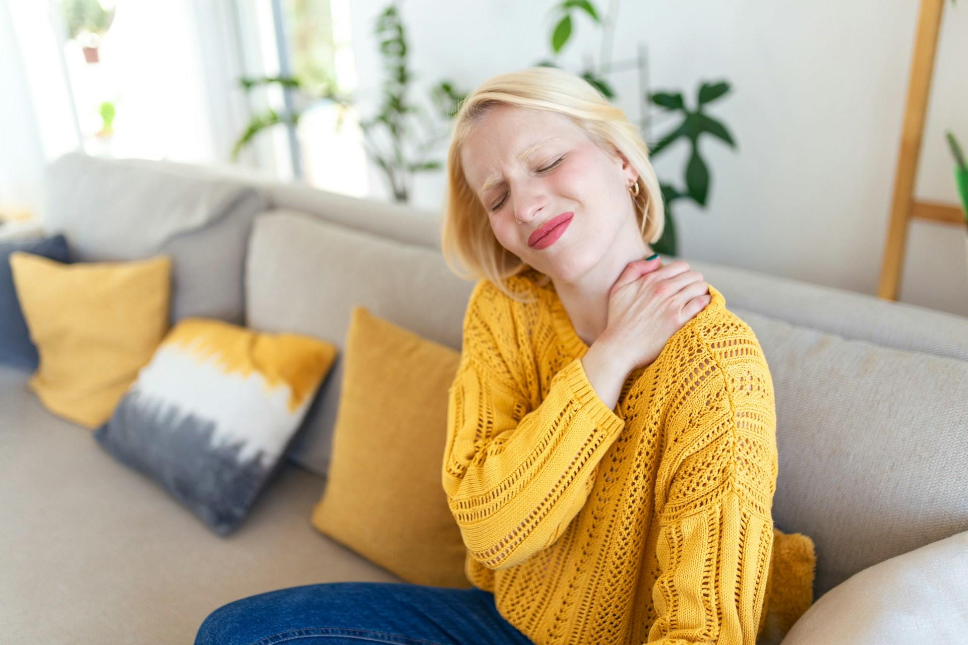 A woman wearing a yellow sweater holds her hand on her shoulder while wincing in pain. 