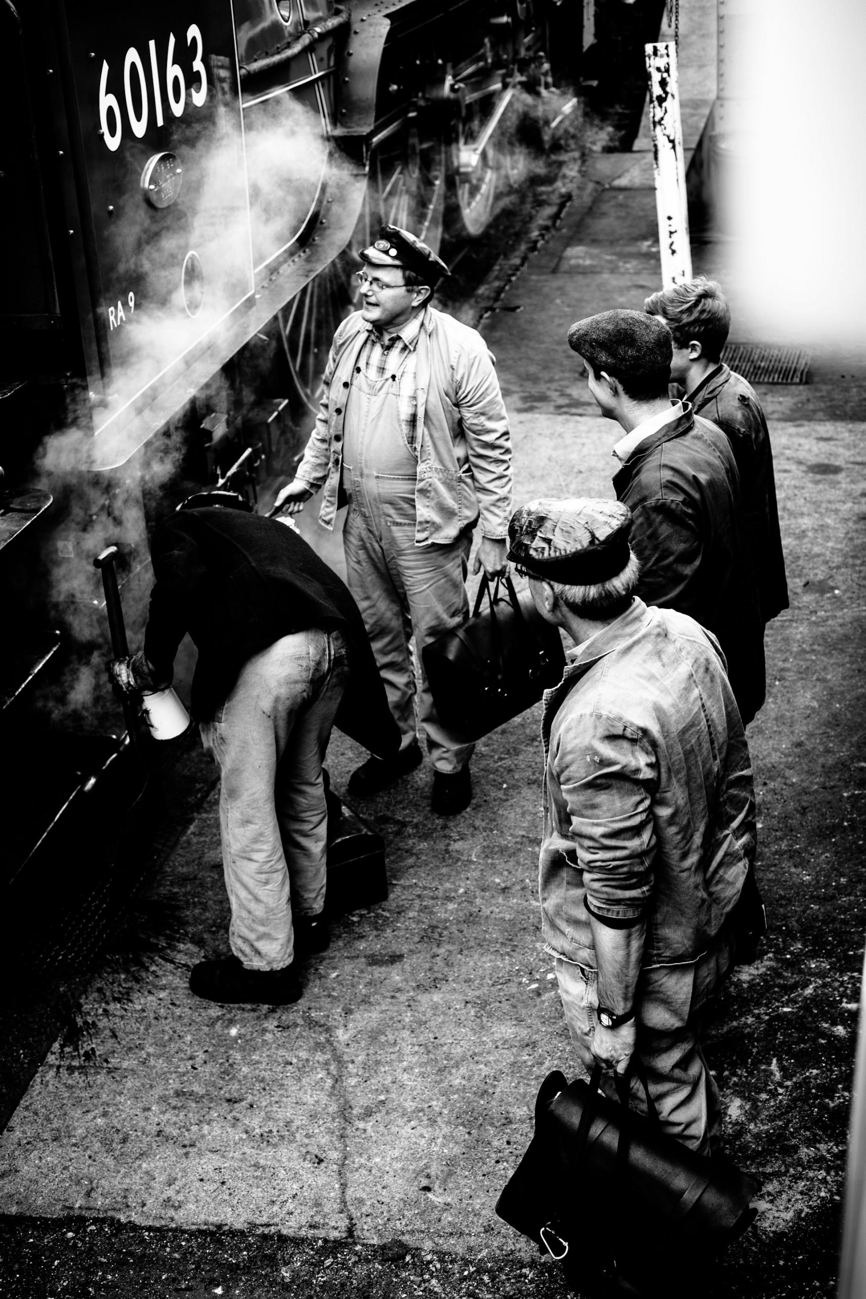A greyscale of men standing by a steaming train. 