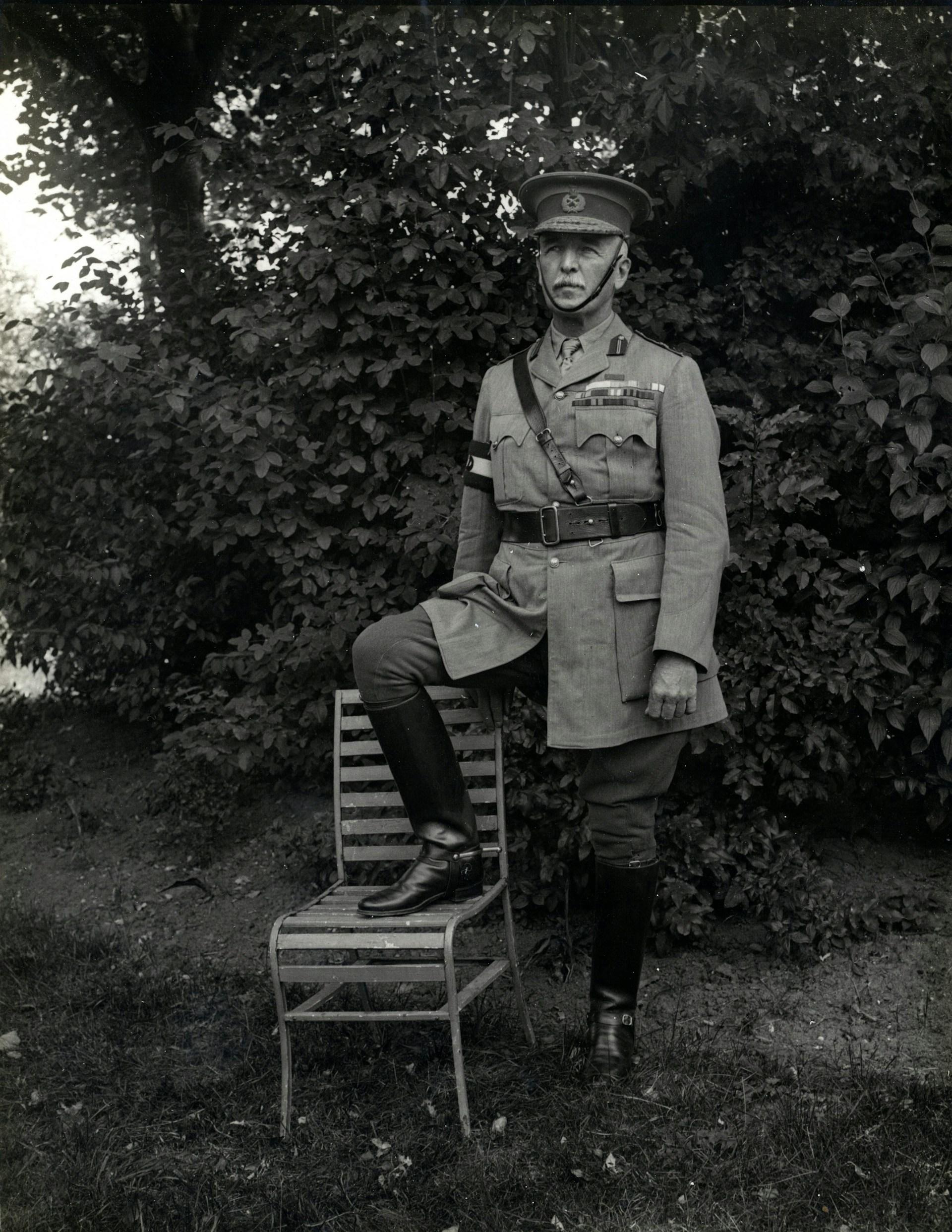 A greyscale of a man wearing a uniform with his foot on a chair, standing outdoors. 