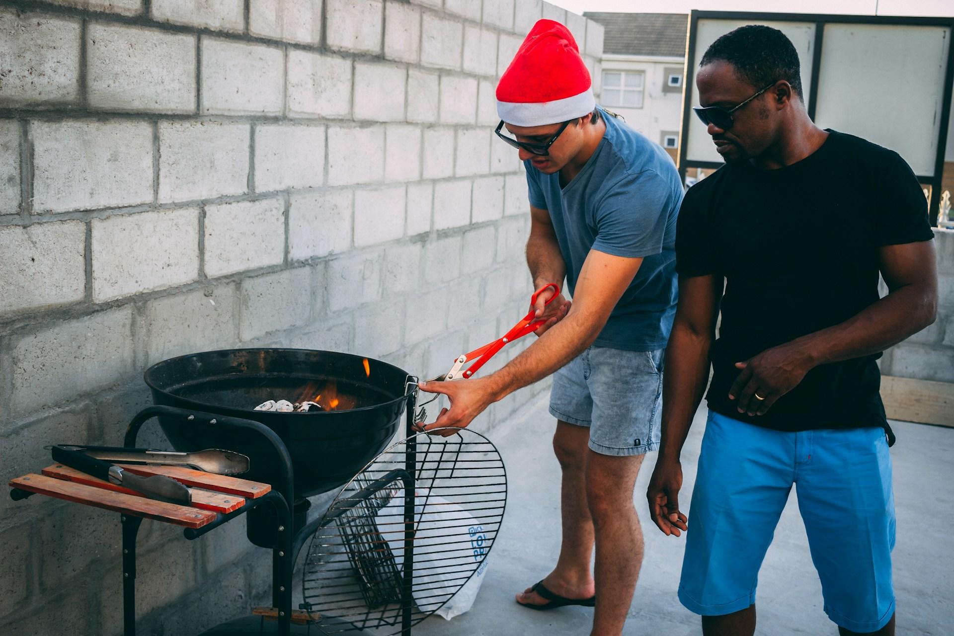 Two men spark up a BBQ, one is wearing a Christmas santa hat.