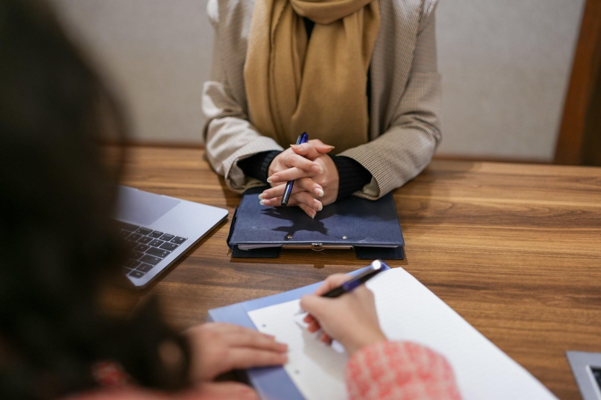 A person wearing a brown jumper holds a pen in their clasped hands, sitting across from a person writing.