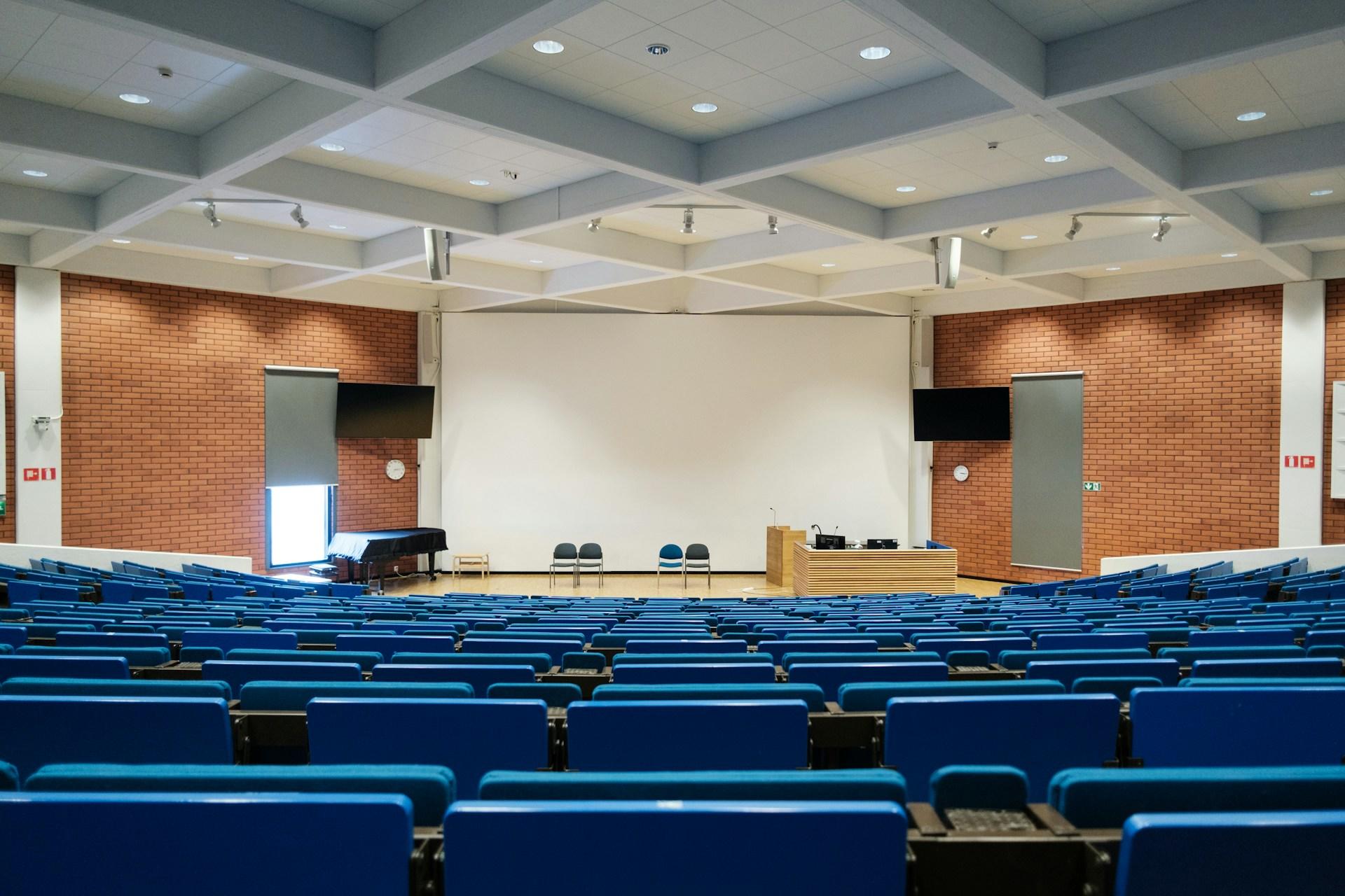 An empty classroom featuring blue seats facing a large screen.