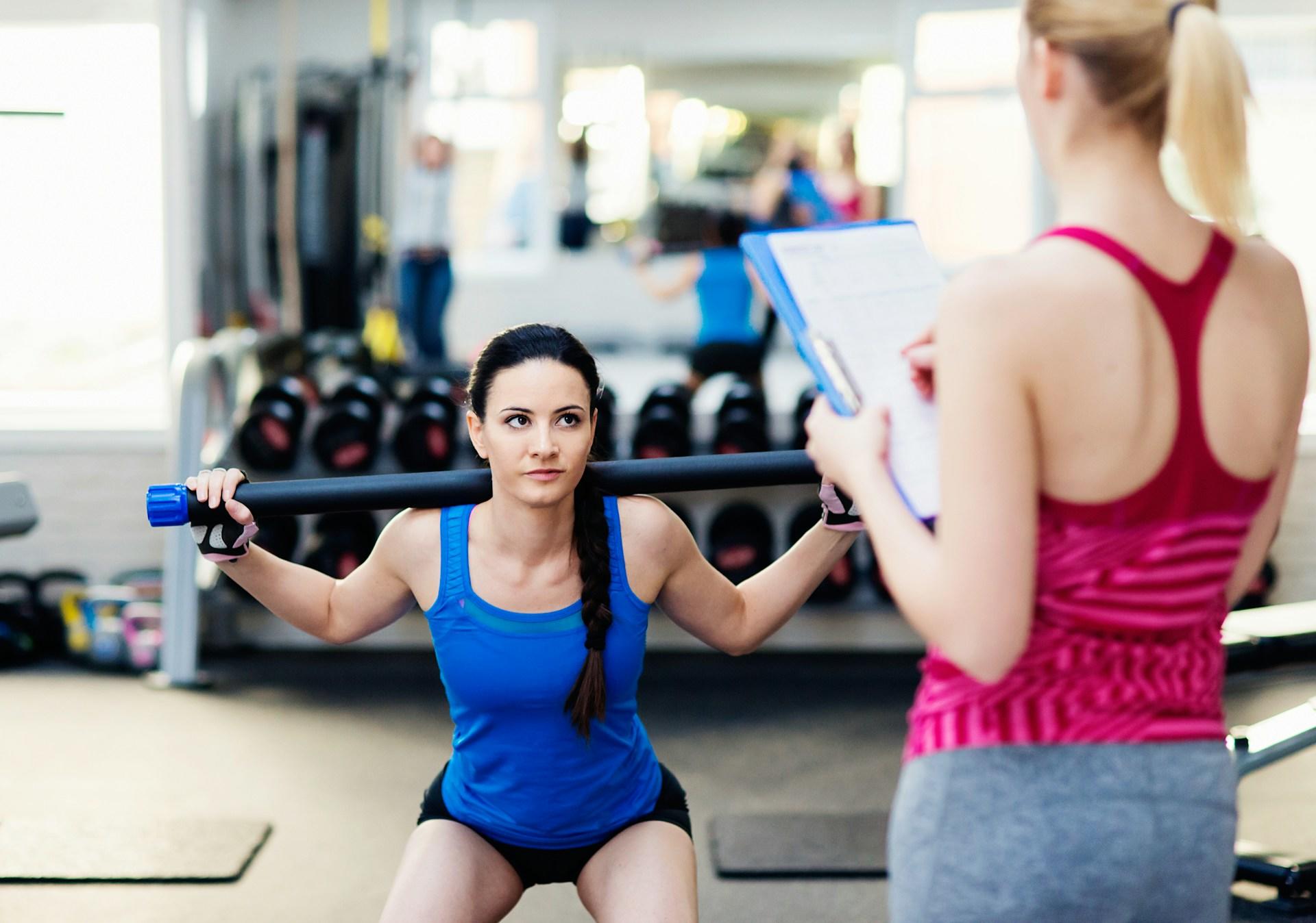A woman with a barbell on her shoulders while another woman watches.