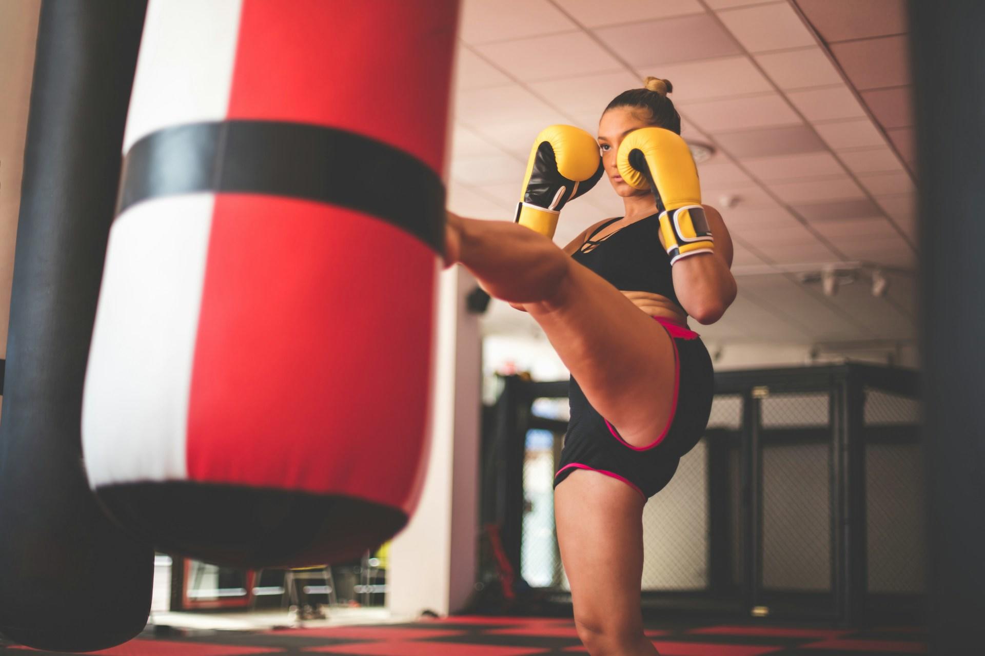 A woman wearing yellow boxing gloves kicks a red bag.