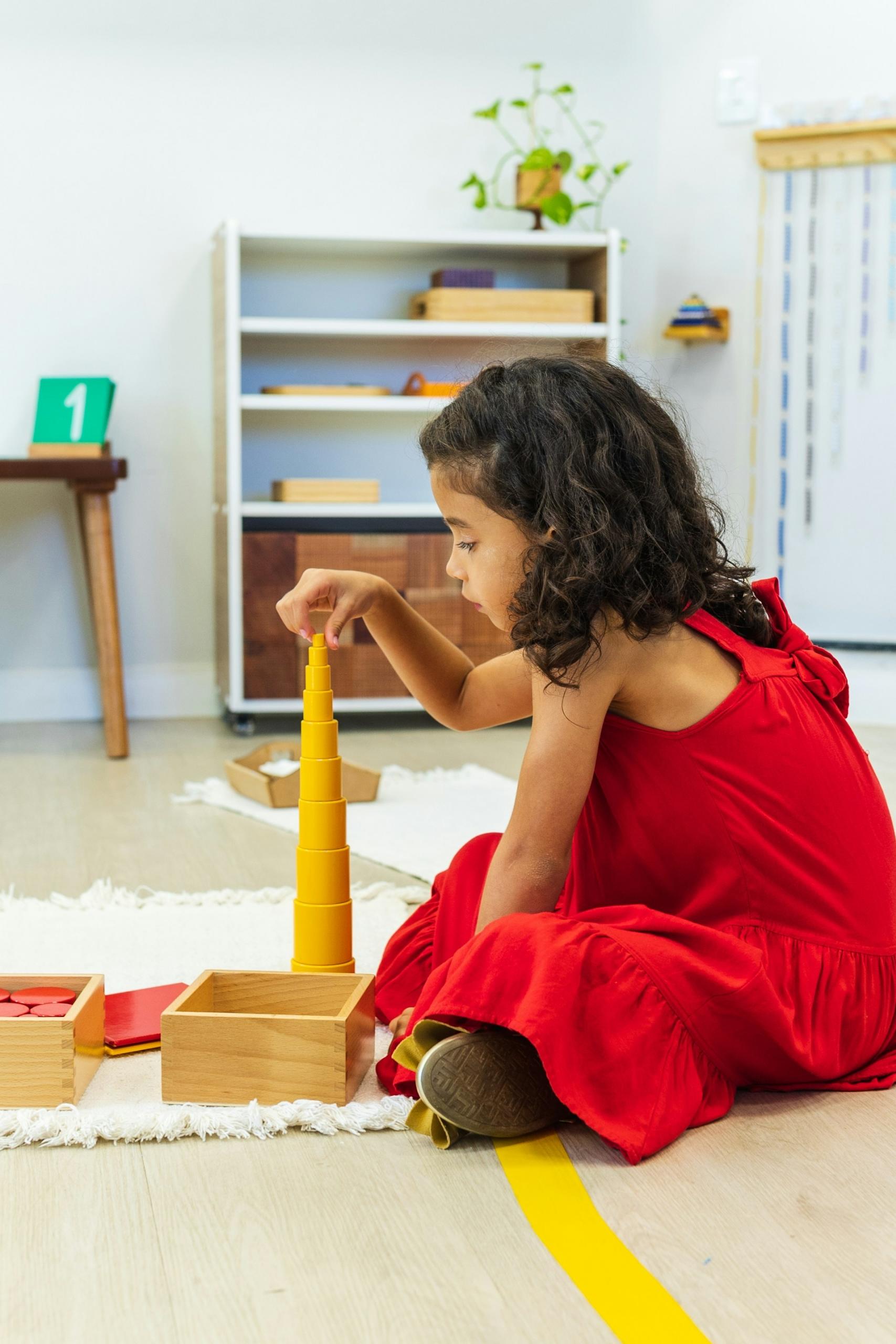 Child in a red dress playing with building blocks on a classroom floor.