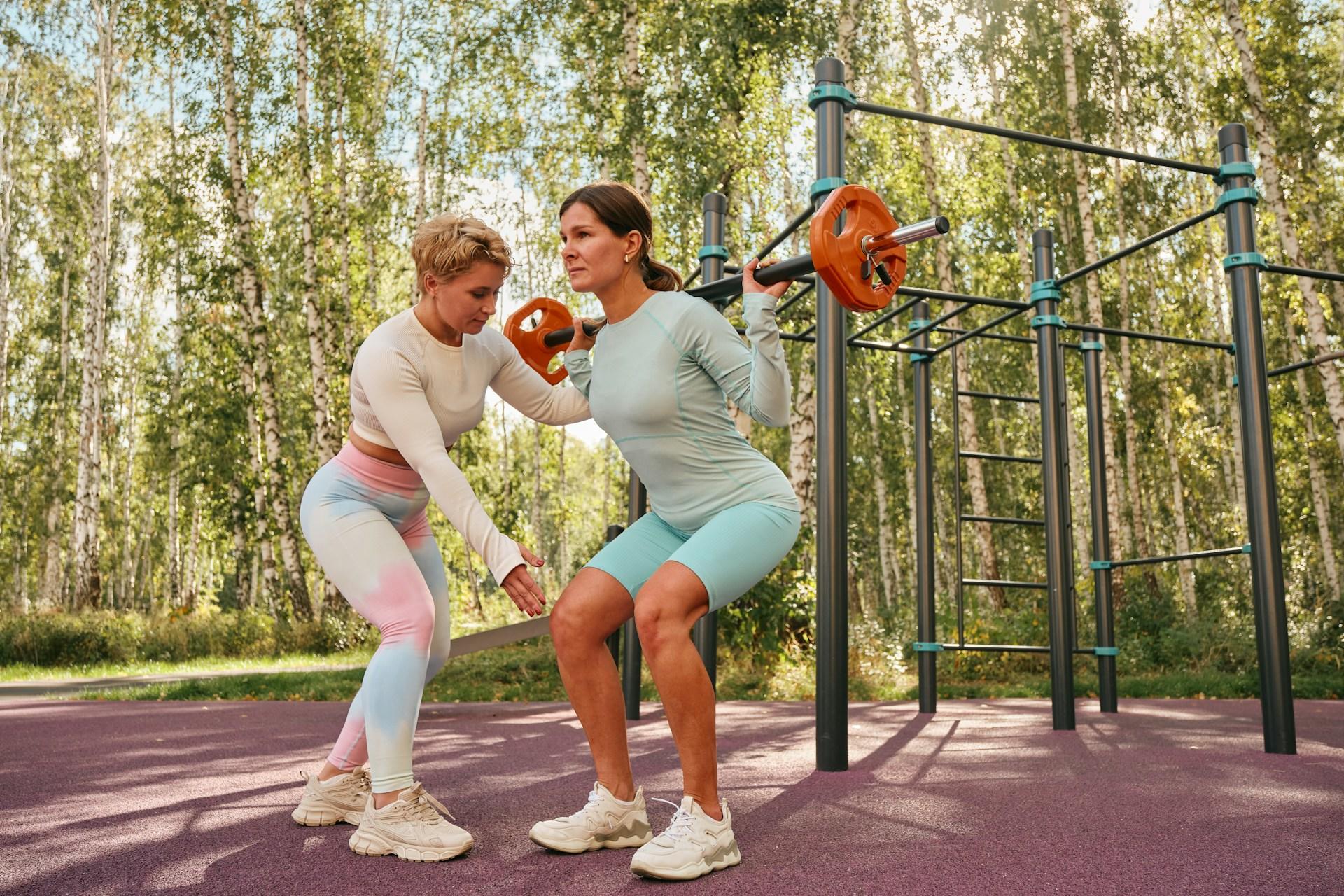 A woman with a barbell on her shoulders next to another guiding the workout.