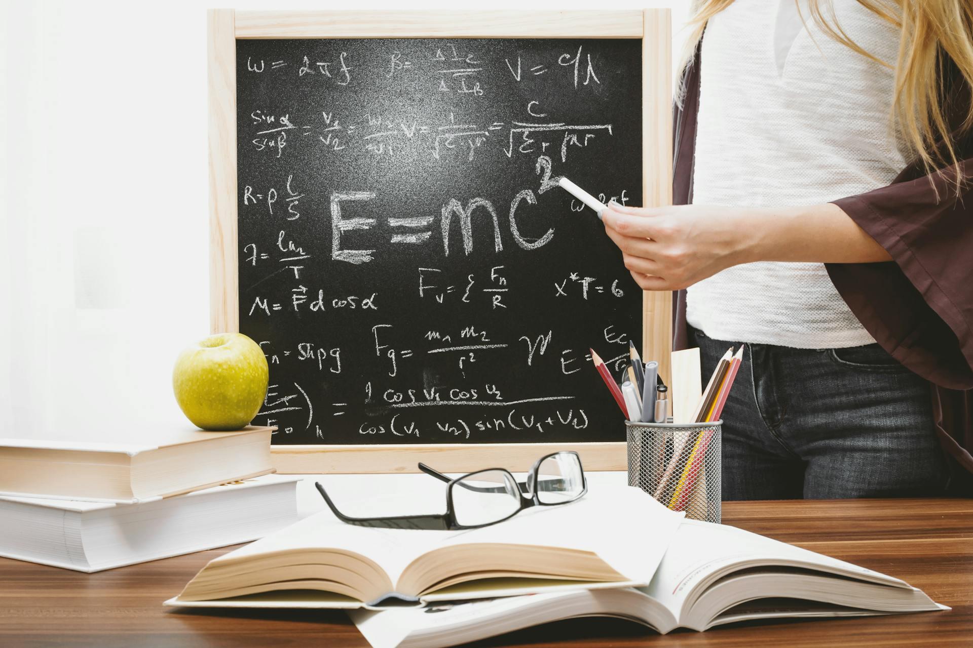 A woman points at a blackboard with physics equations on it.