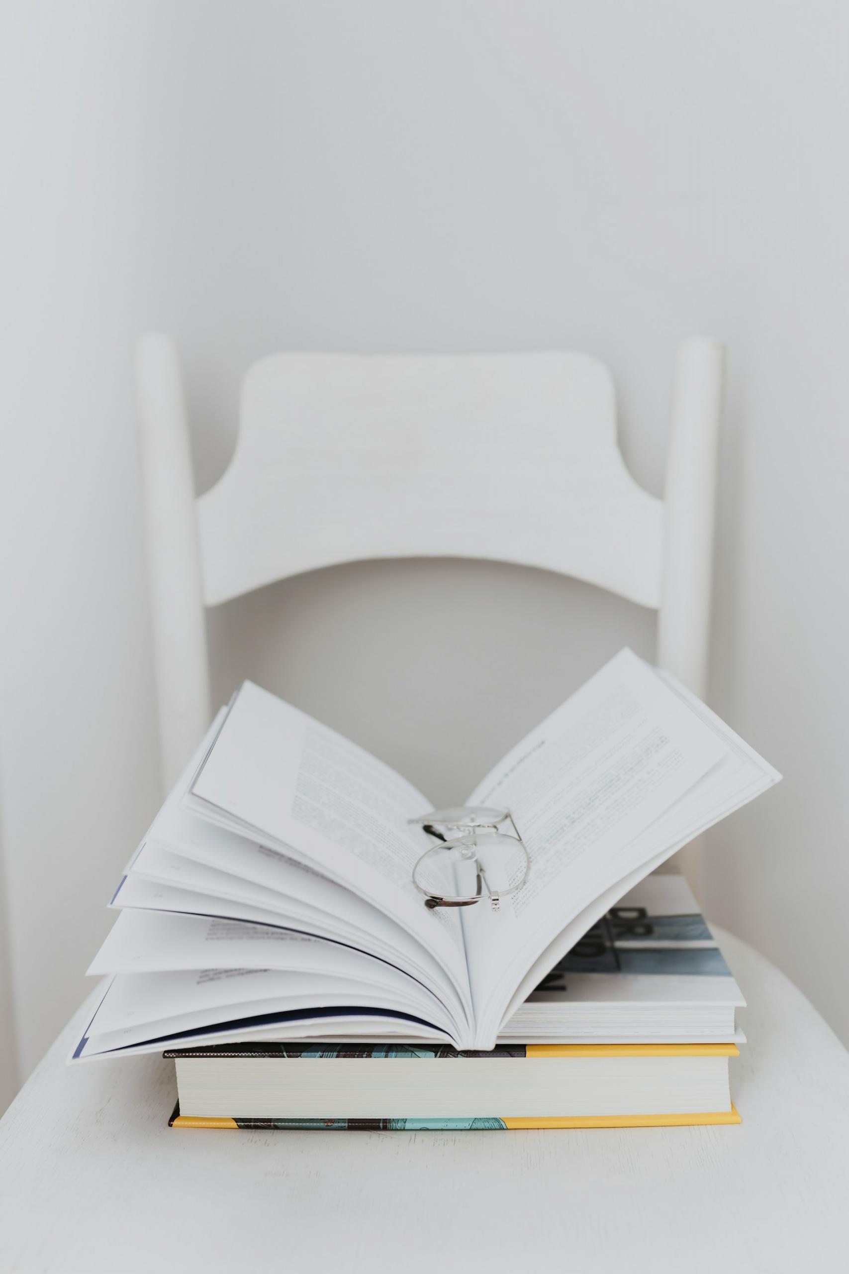 An open book stacked on two others, on a white chair.