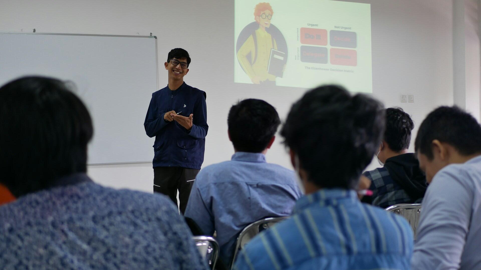 A person wearing blue standing in front of a classroom narrating a slideshow.