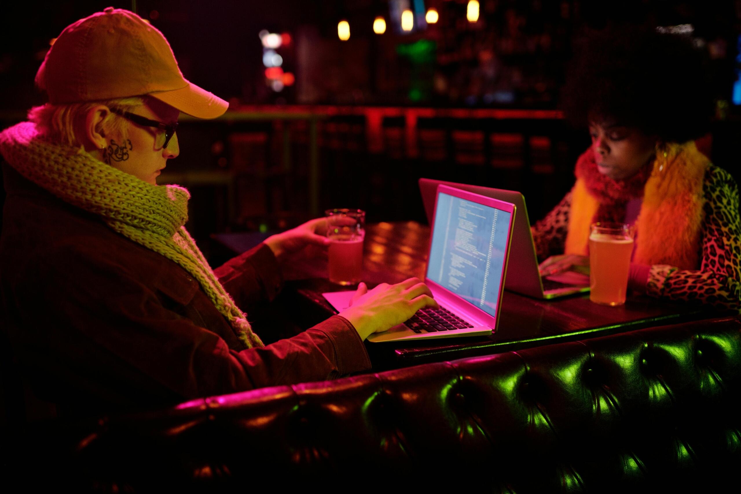 A man and a woman sitting in a bar, both using their laptops with lines of code on the screens.