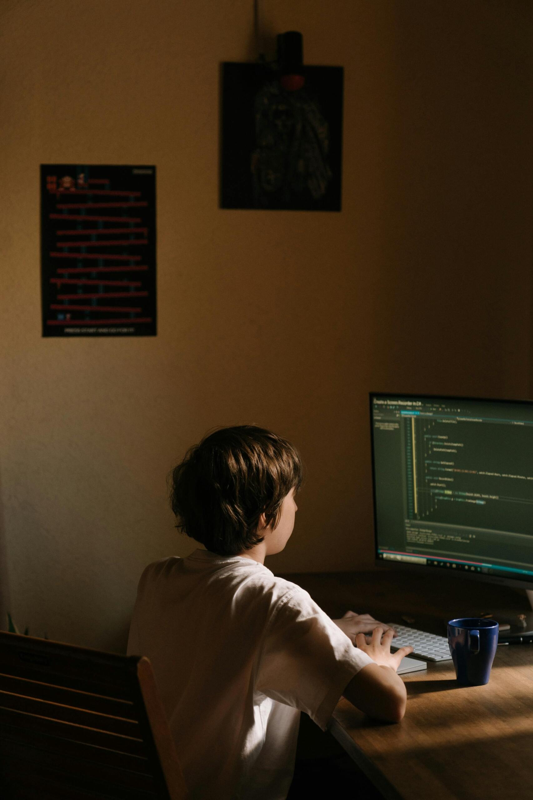 A young boy in a white shirt sitting on a chair, focused on coding on his computer.