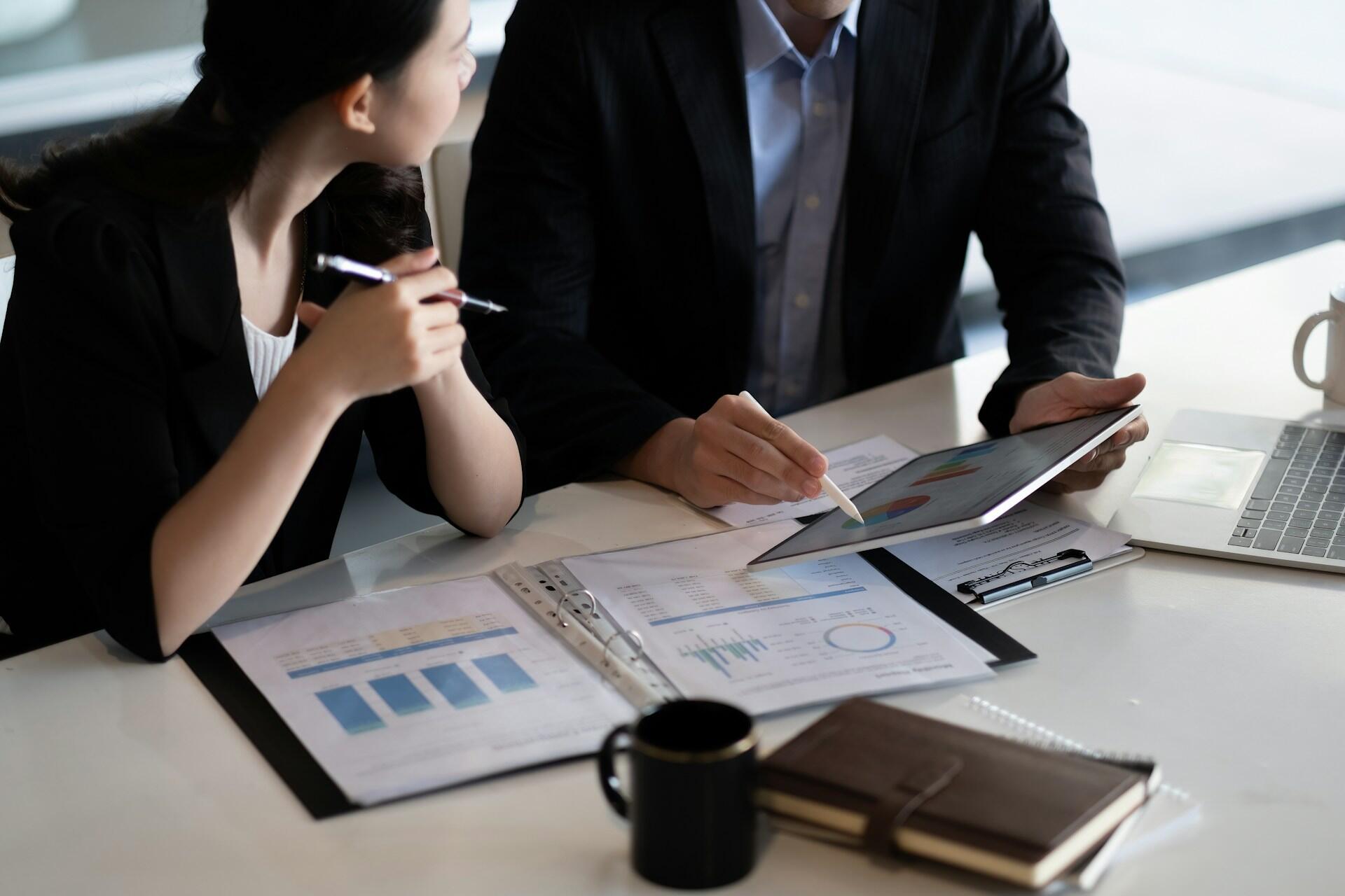 Two business professionals seated at a table, engaged with a laptop and documents in a collaborative setting.