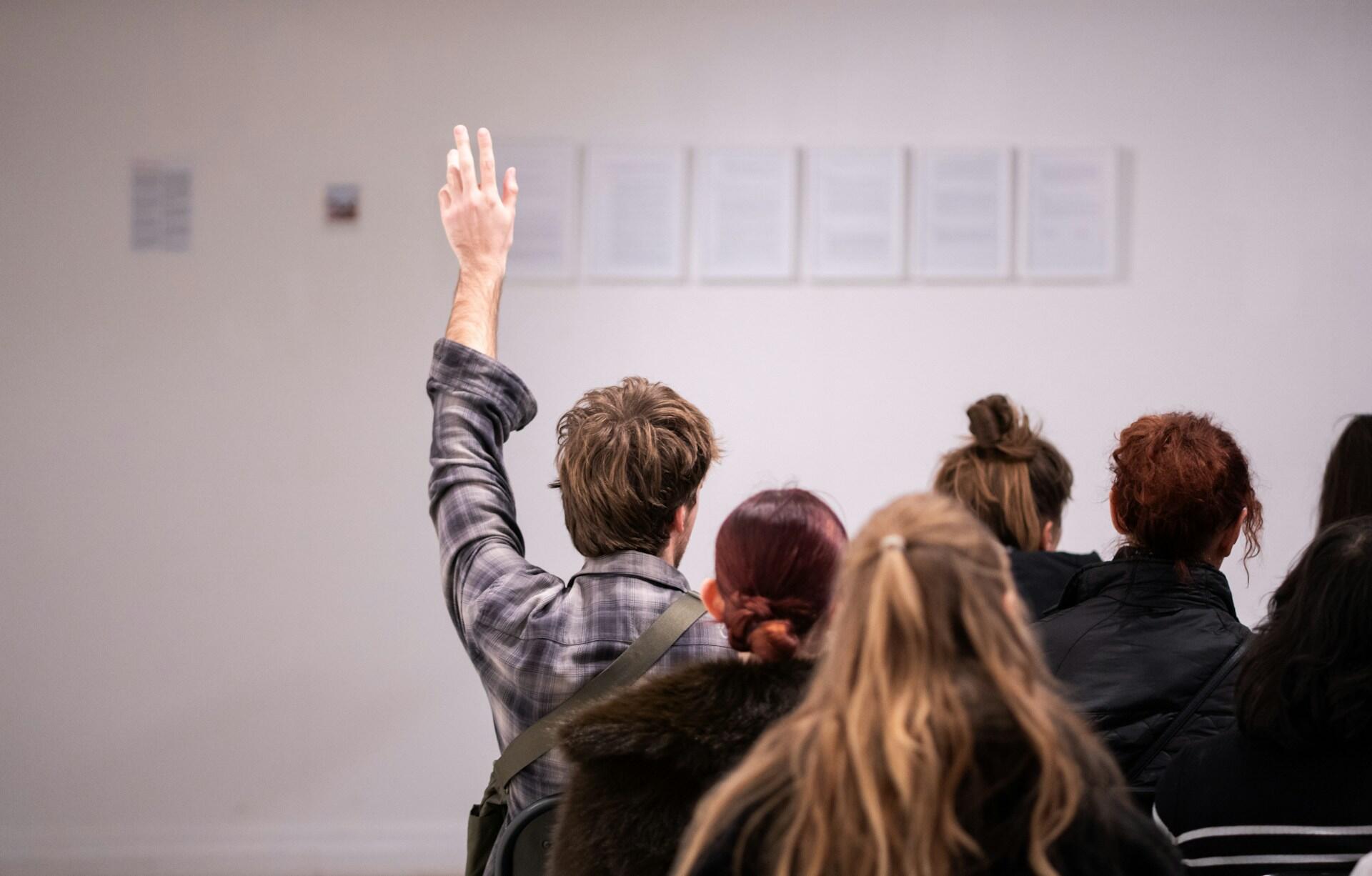 A person raising their hand among a seated group facing a white wall with framed papers or artworks.